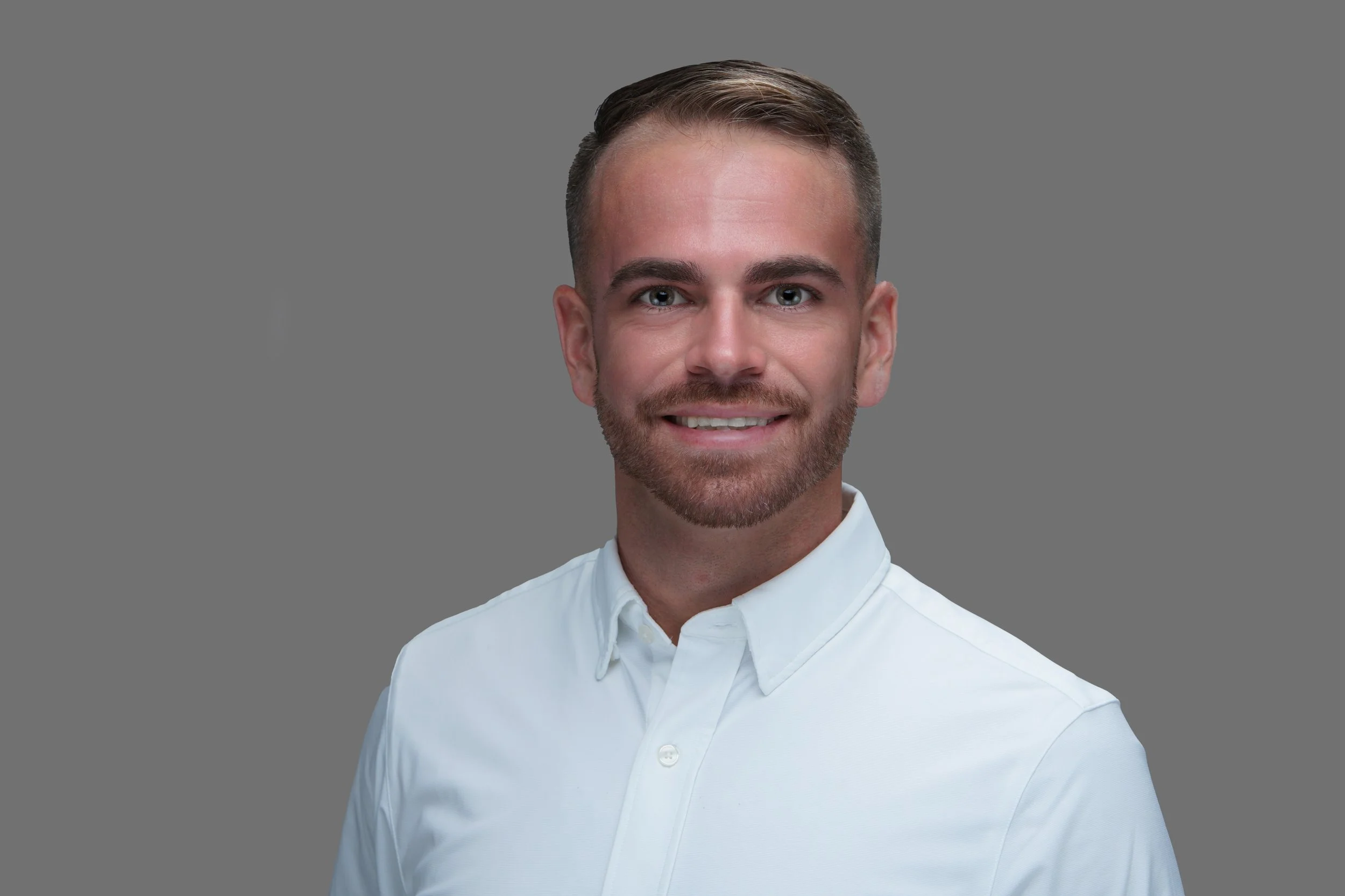 A young man with light skin, brown hair, and a trimmed beard, smiling and wearing a white collared shirt, standing against a plain gray background.