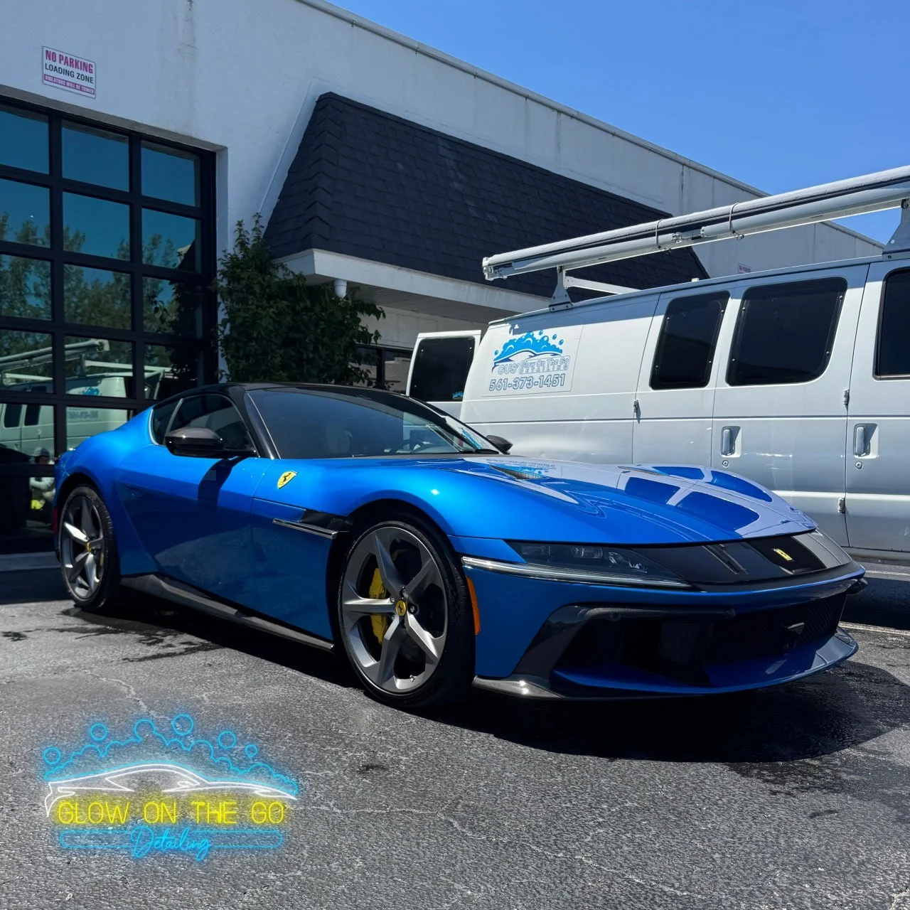 A blue Ferrari sports car parked outside a building with a white van nearby. The car has black and yellow details, including Ferrari logos and yellow brake calipers. The building has large glass windows and a no parking sign visible on the wall.
