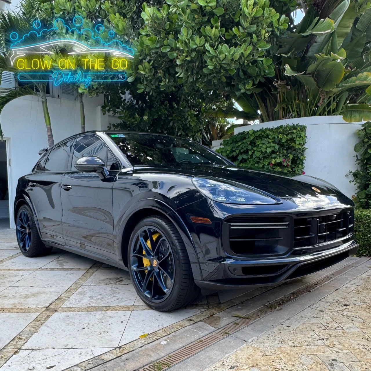 Black Porsche SUV parked on a stone driveway with lush green trees and bushes in the background, and a neon sign that reads 'Glow on the Go Detailing' hanging above.