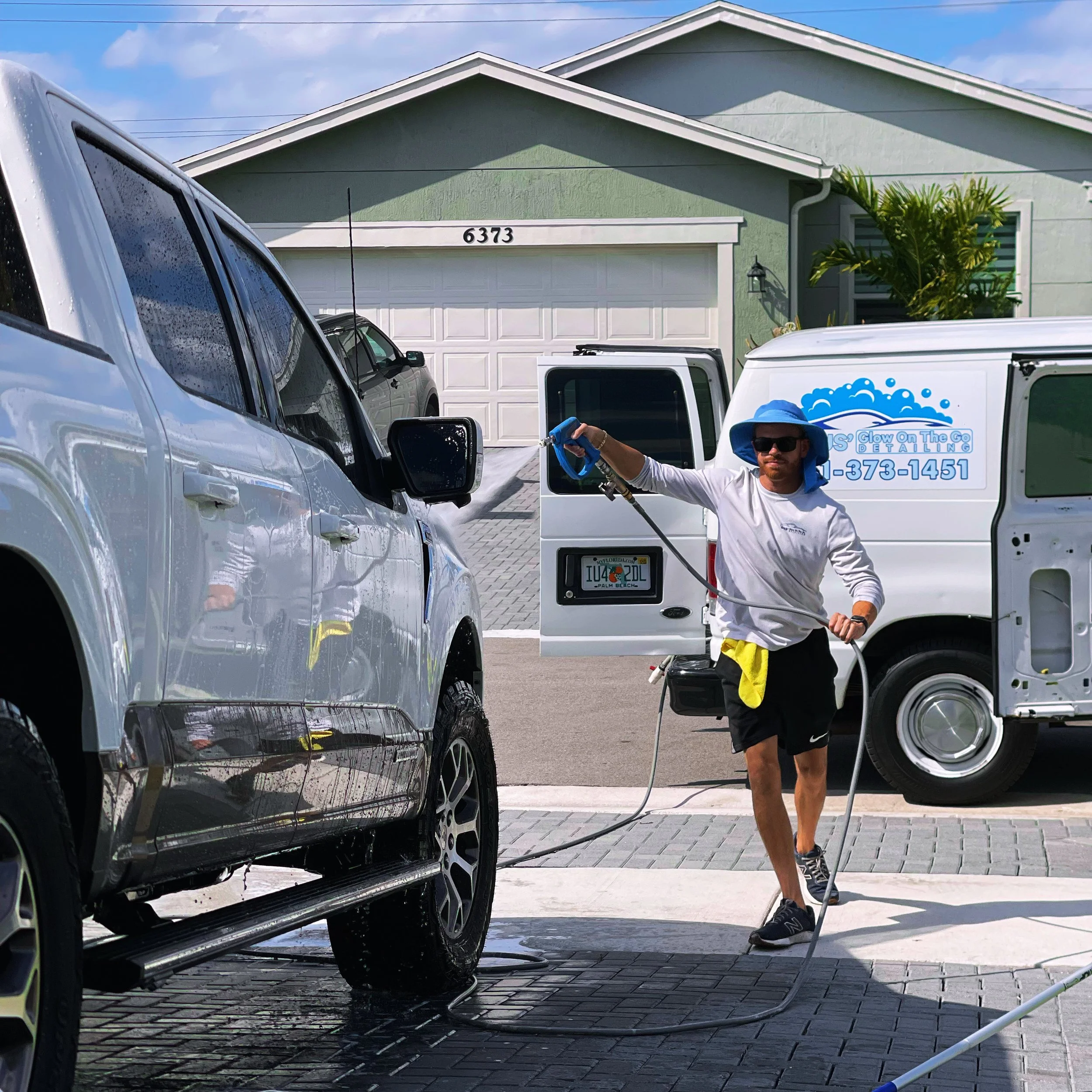 A man in casual clothes, wearing sunglasses and a wide-brimmed hat, is pressure washing a black car with a hose and spray nozzle. The car is parked on a driveway in front of a green house with white trim and a garage door. Several other vehicles are also in the driveway, including a white van with a logo and phone number, and an SUV. The sky is partly cloudy.