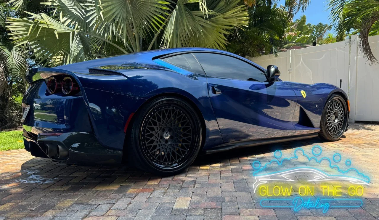 A blue Ferrari sports car parked on a brick driveway with lush green palm trees and a white fence in the background. There is a neon sign in the lower right corner that reads 'Glow on the Go Detailing'.