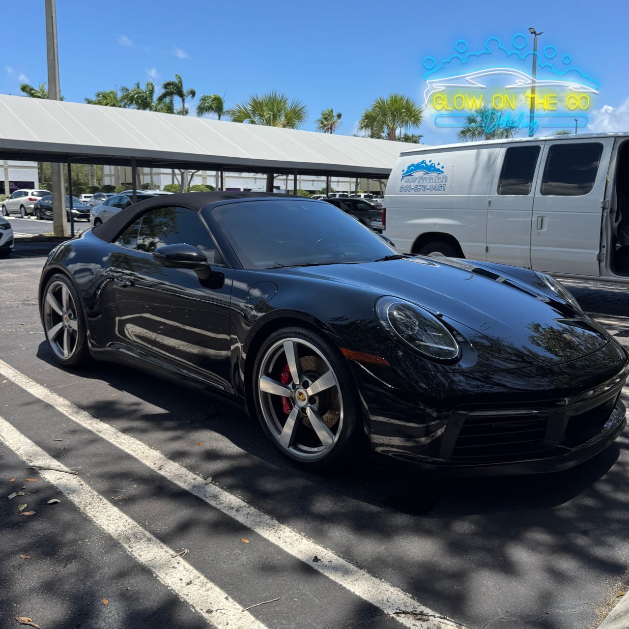 A black Porsche convertible sports car parked in a lot under a partly cloudy sky with shadows of trees on the pavement. There are other cars and palm trees in the background, with a vehicle repair shop and a neon sign reading 'GLOW ON THE GO'.