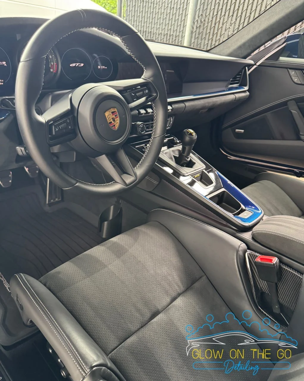 Interior of a Porsche GT3 sports car featuring a black leather steering wheel with the Porsche crest, digital instrument cluster, manual gear shifter, and black seats. The dashboard is sleek and modern with minimal buttons.