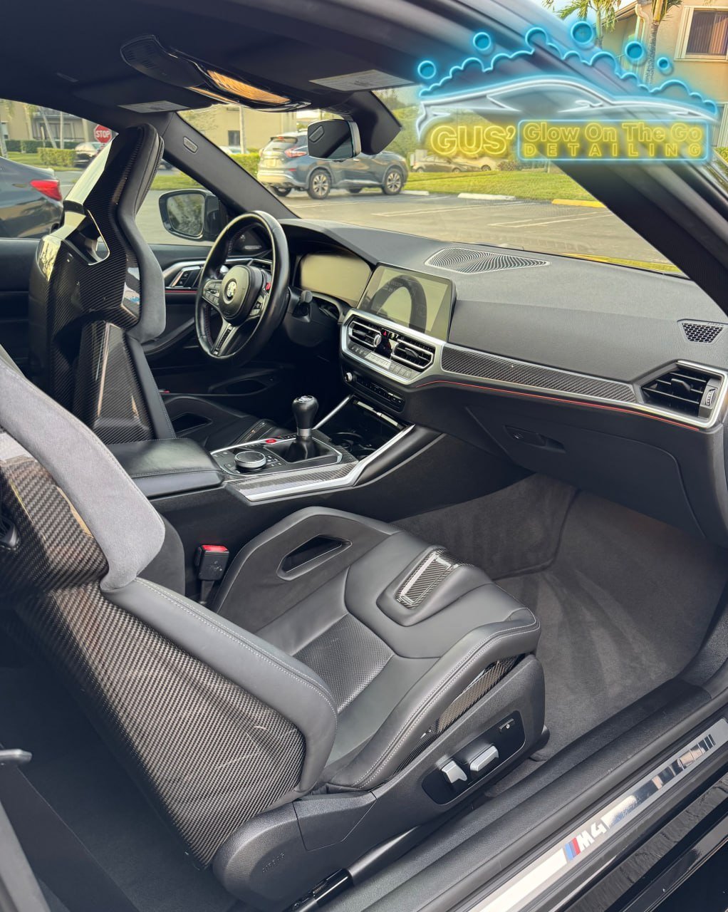 The interior of a luxury sports car, showing carbon fiber accents, a digital dashboard, gearshift, racing seats with carbon fiber shells, and a neon sign on the windshield reading 'GUS' Glow On The Go Detailing' with a stylized car outline.