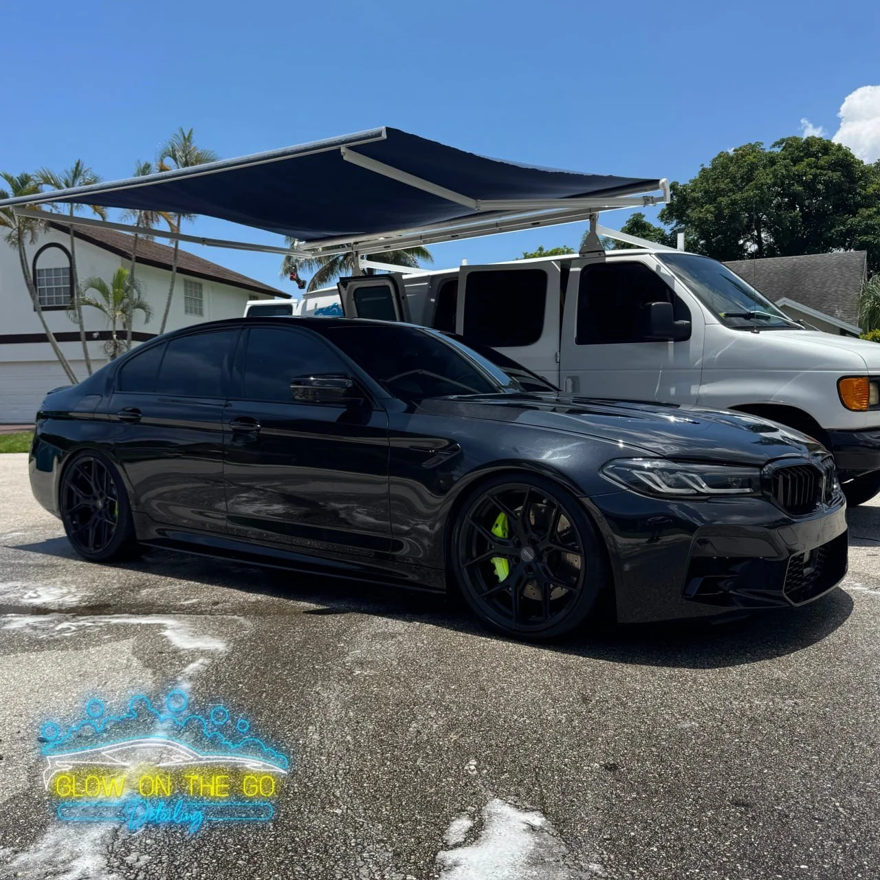 A black BMW sports car with tinted windows parked next to a white van with an attached blue awning. The ground appears wet, and a neon sign on the pavement reads 'Glow on the Go detailing' with a car illustration and bubbles.
