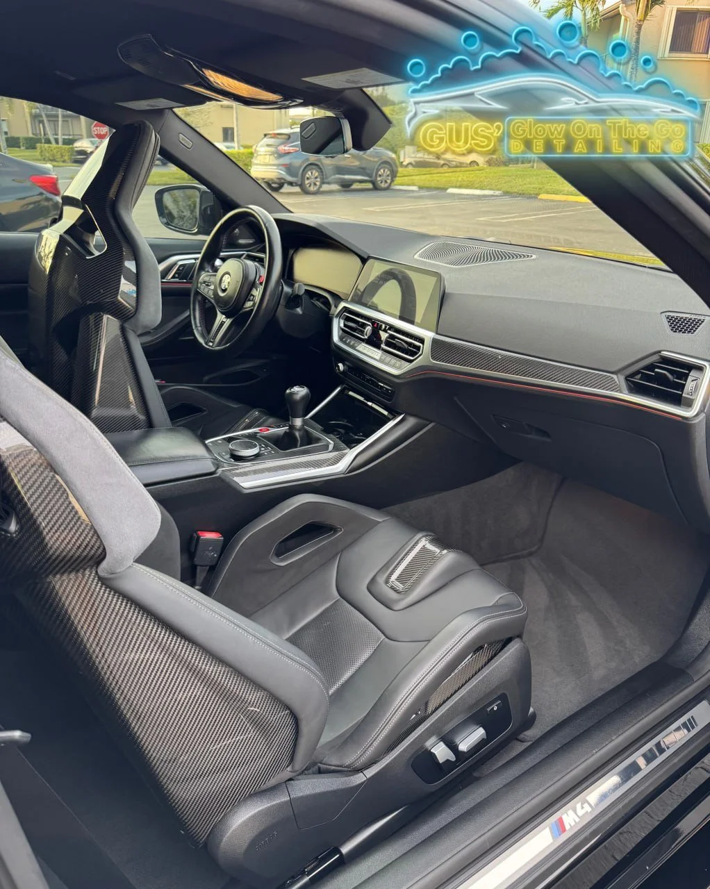 The interior of a luxury car with carbon fiber accents, a digital dashboard, leather seats, and a neon sign in the window that reads 'GUS Glow On The Go Detailing'.
