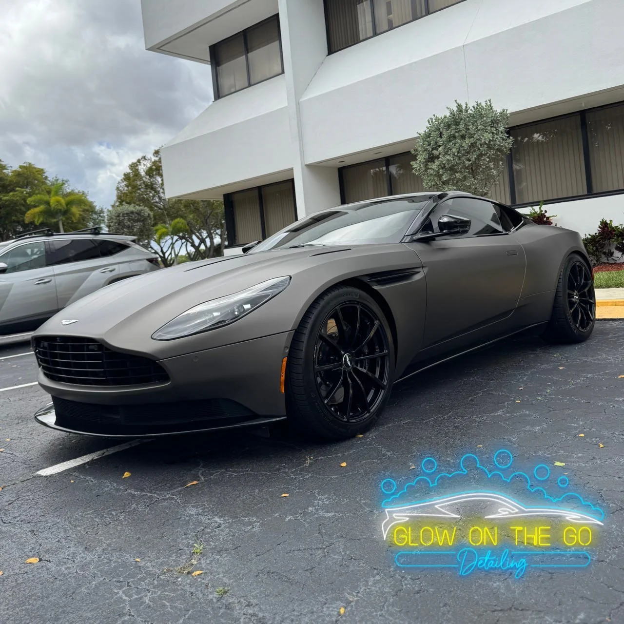 A gray Aston Martin sports car parked in an outdoor lot under cloudy skies, with black alloy wheels and tinted windows, next to other vehicles and modern buildings.