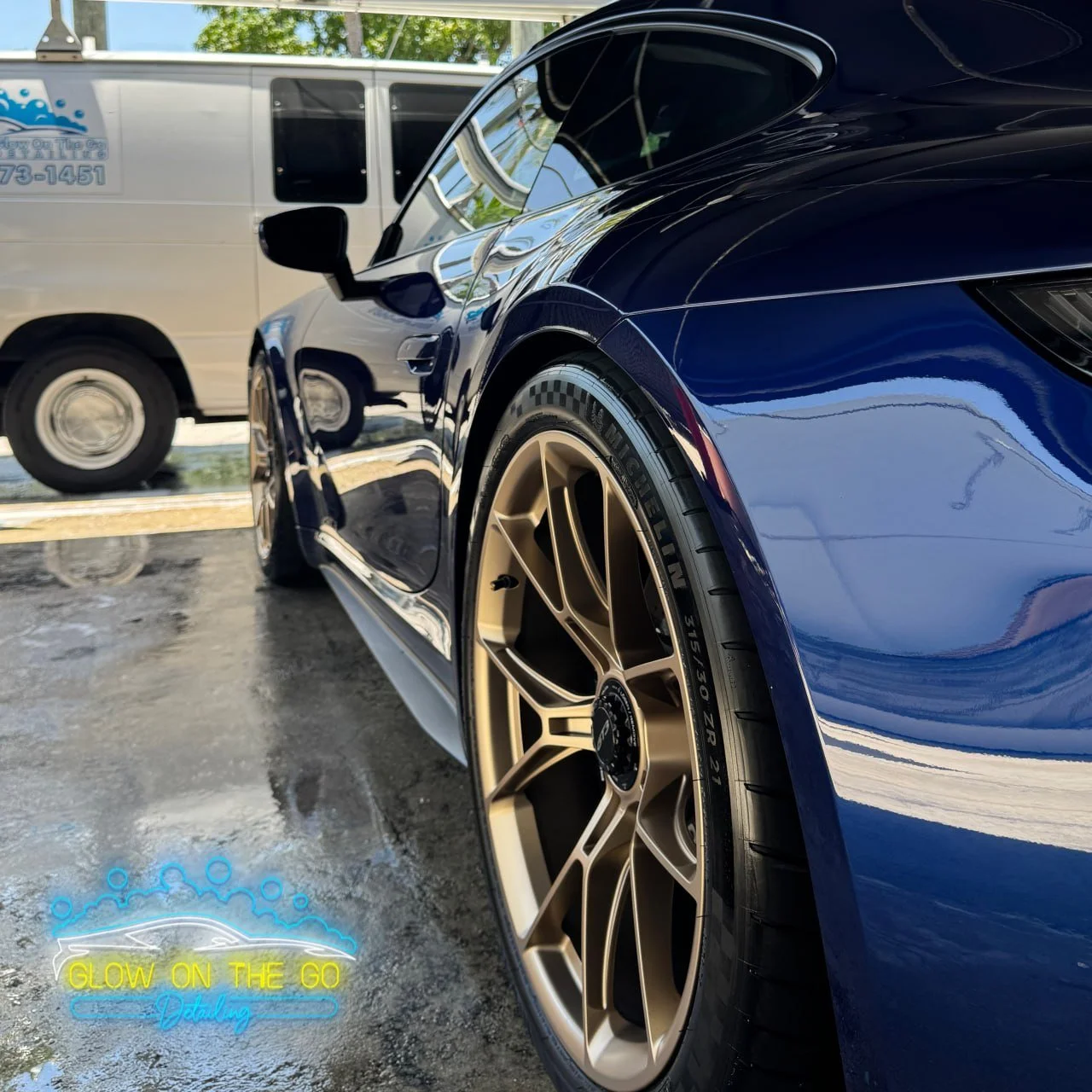 Close-up of a shiny navy blue sports car with gold wheels parked on a wet surface, with a white van in the background.