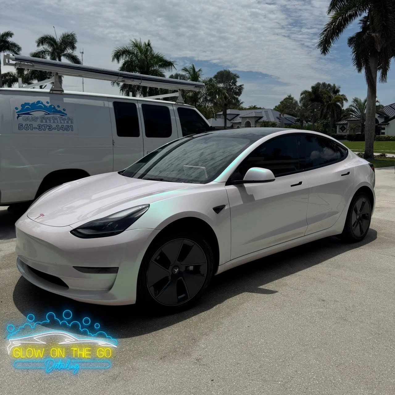 A white Tesla Model 3 parked on a driveway with a white service van and trees in the background under a cloudy sky.