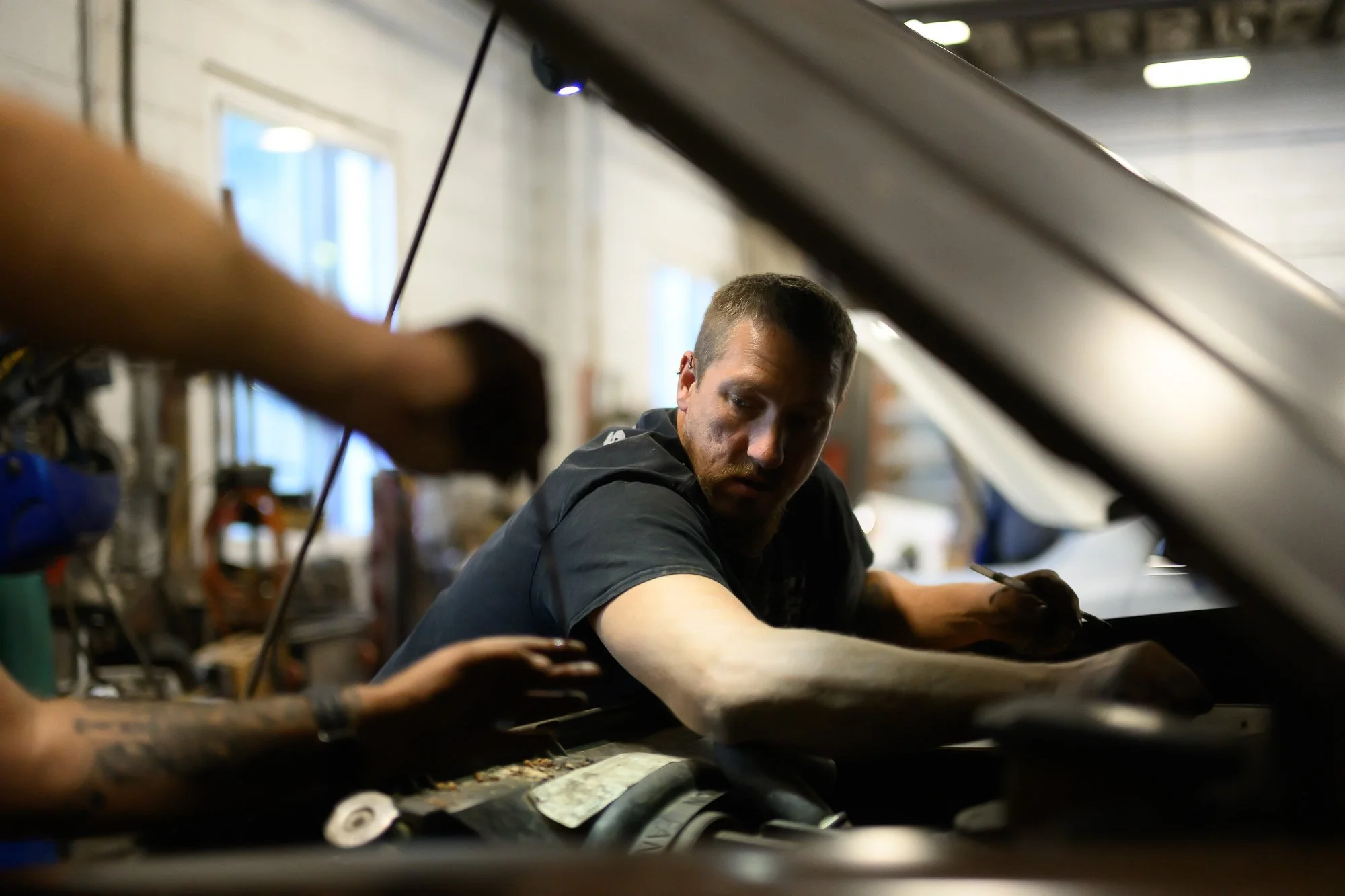 A man working on a vehicle in a garage or workshop. He is leaning over the engine bay, focused on his work, with tools and equipment visible in the background.