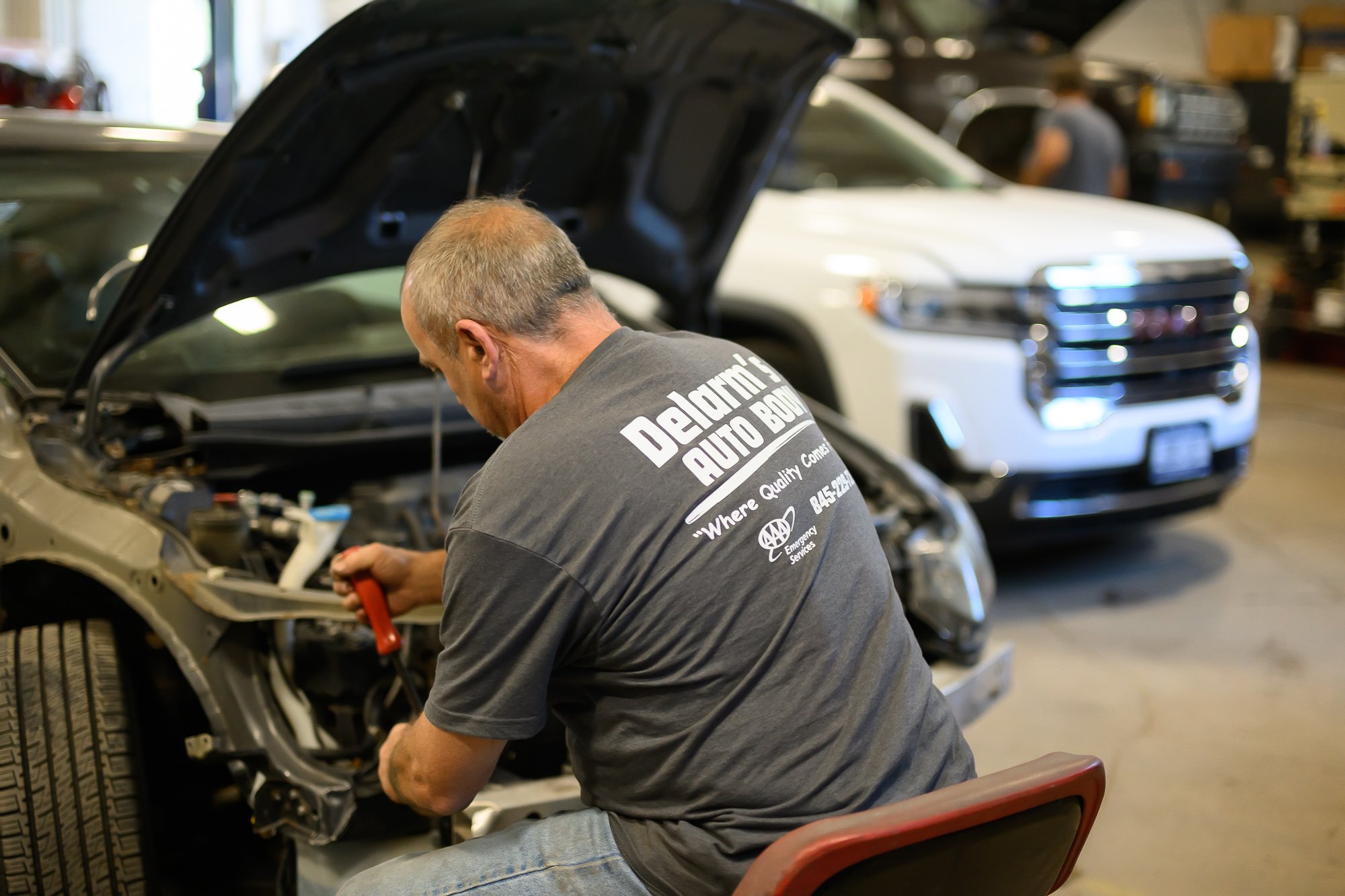 A mechanic is working on a car in a garage, with the hood open. The mechanic wears a gray shirt with white text that reads 'Delarthe's Auto Body' and is using a tool on the vehicle. In the background, trucks are parked inside the garage.