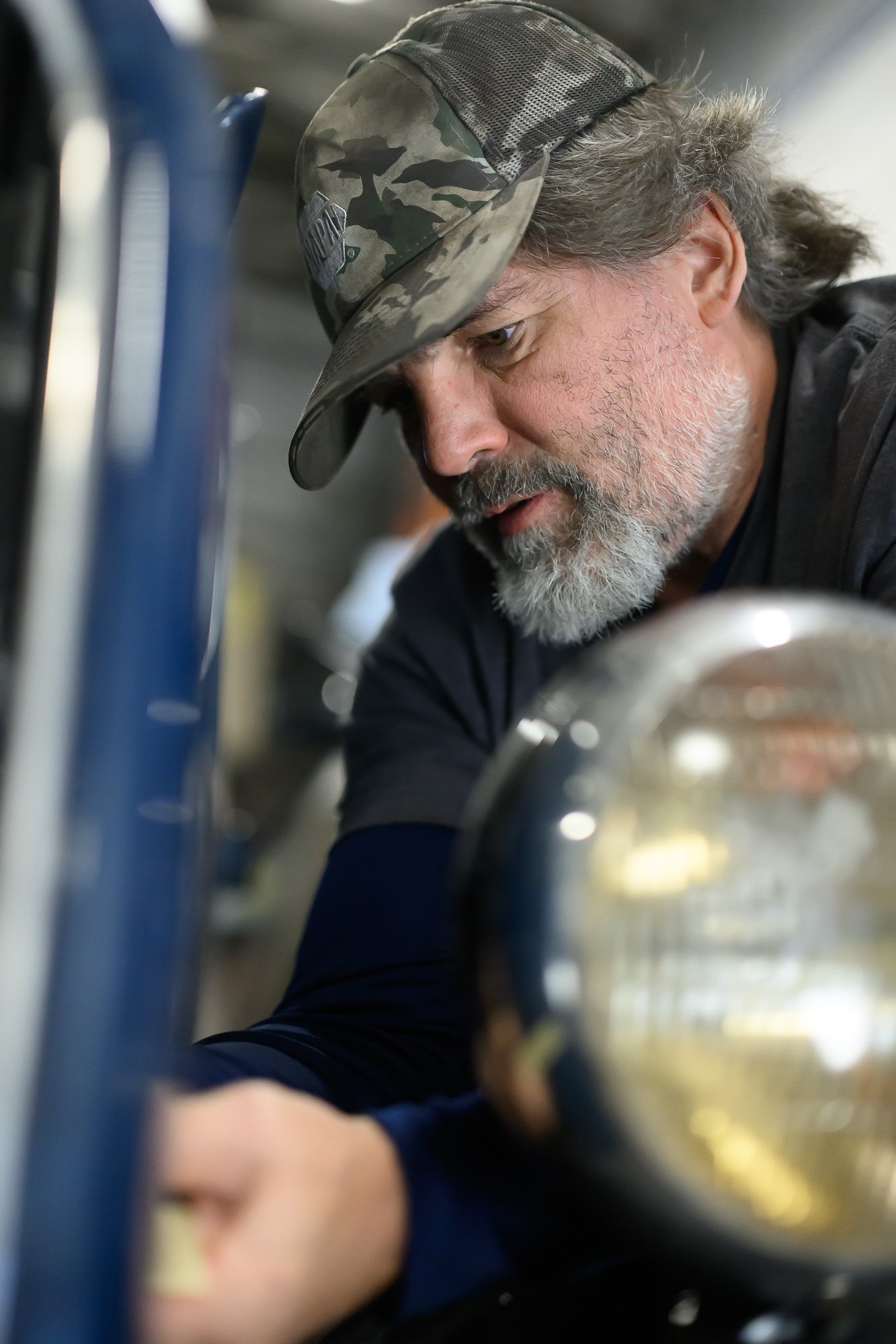 Close-up photo of a man wearing a camouflage baseball cap looking down, with a gray beard and long hair, in a setting with shiny metallic and blue parts.