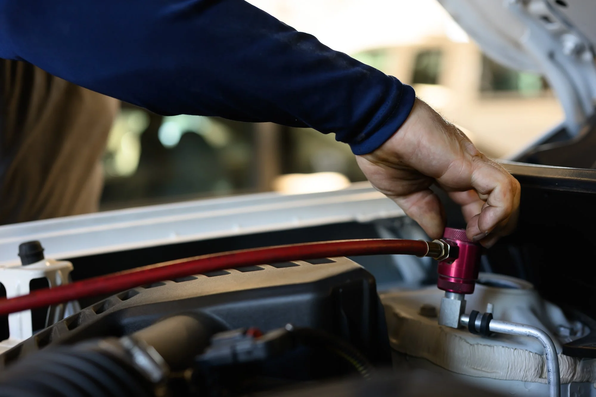 A person wearing a dark blue long sleeve shirt working on a mechanical or automotive component, using their hand to connect or adjust a pink fitting on a red and black hose.