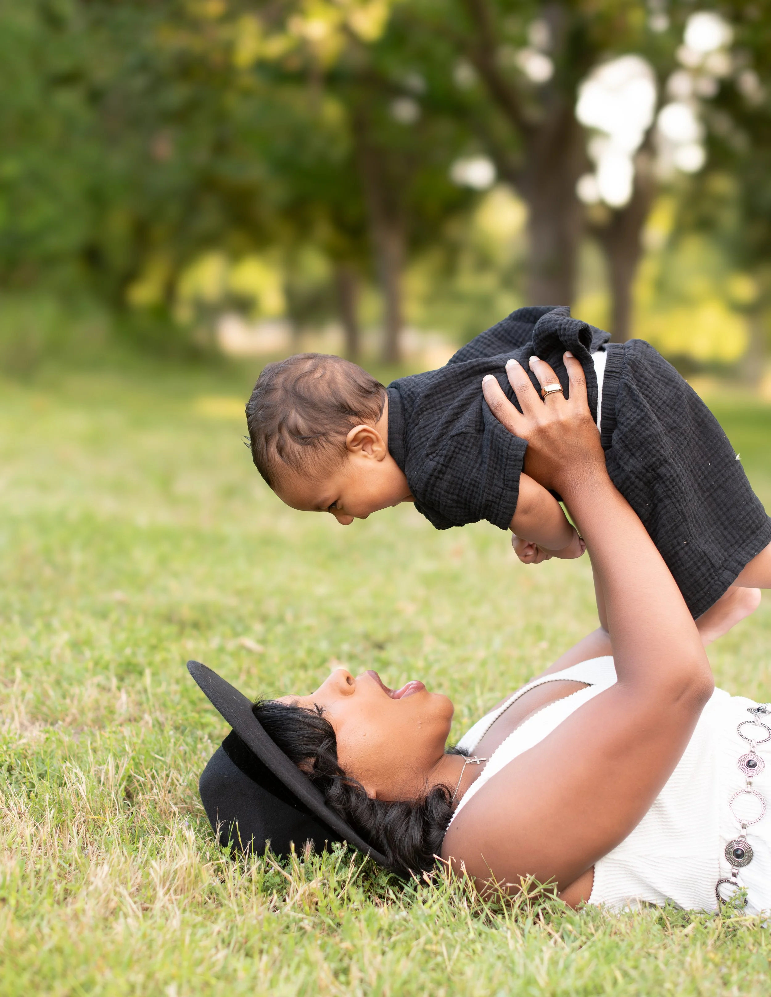 A woman lying on the grass in a park, facing an upside-down child who is on her lap. The woman is wearing a white sleeveless top and a black hat, while the child is wearing a black jacket. The woman has her hands on the child's back, and they are both engaged in a playful moment outdoors with trees in the background.