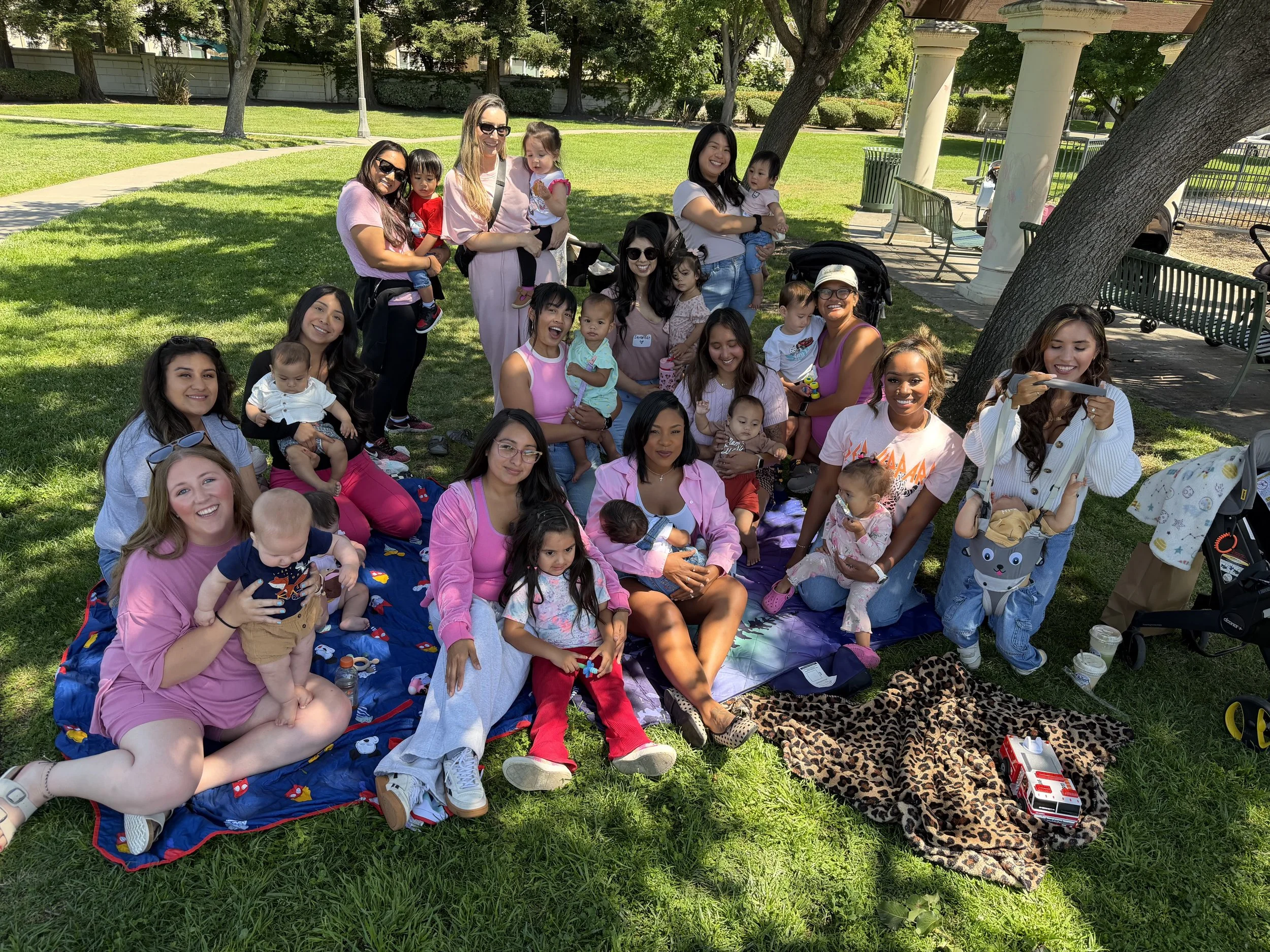 Group of women and children gathered outdoors under a tree in a park, sitting on blankets and benches, smiling and taking photos, with toys and drinks nearby.
