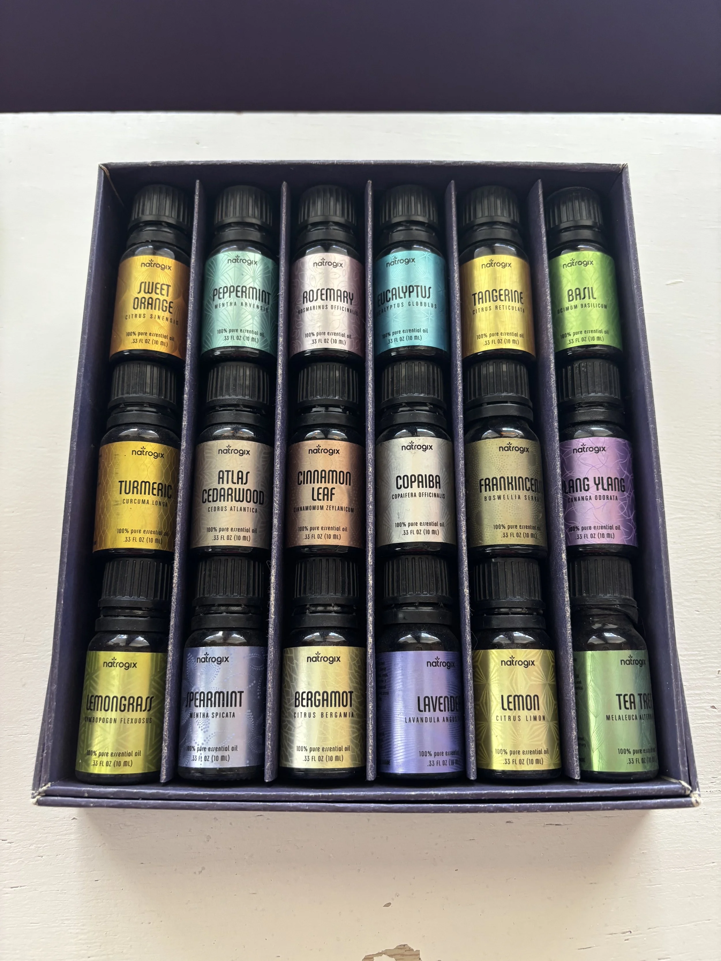 A box of essential oils with multiple bottles arranged in rows. Each bottle has a colorful label with the name of the oil, such as Sweet Orange, Peppermint, Rosemary, Eucalyptus, Tangerine, Basil, Turmeric, Cedarwood, Cinnamon Leaf, Copaiba, Frankincense, Lavender, Lemon, Tea Tree.