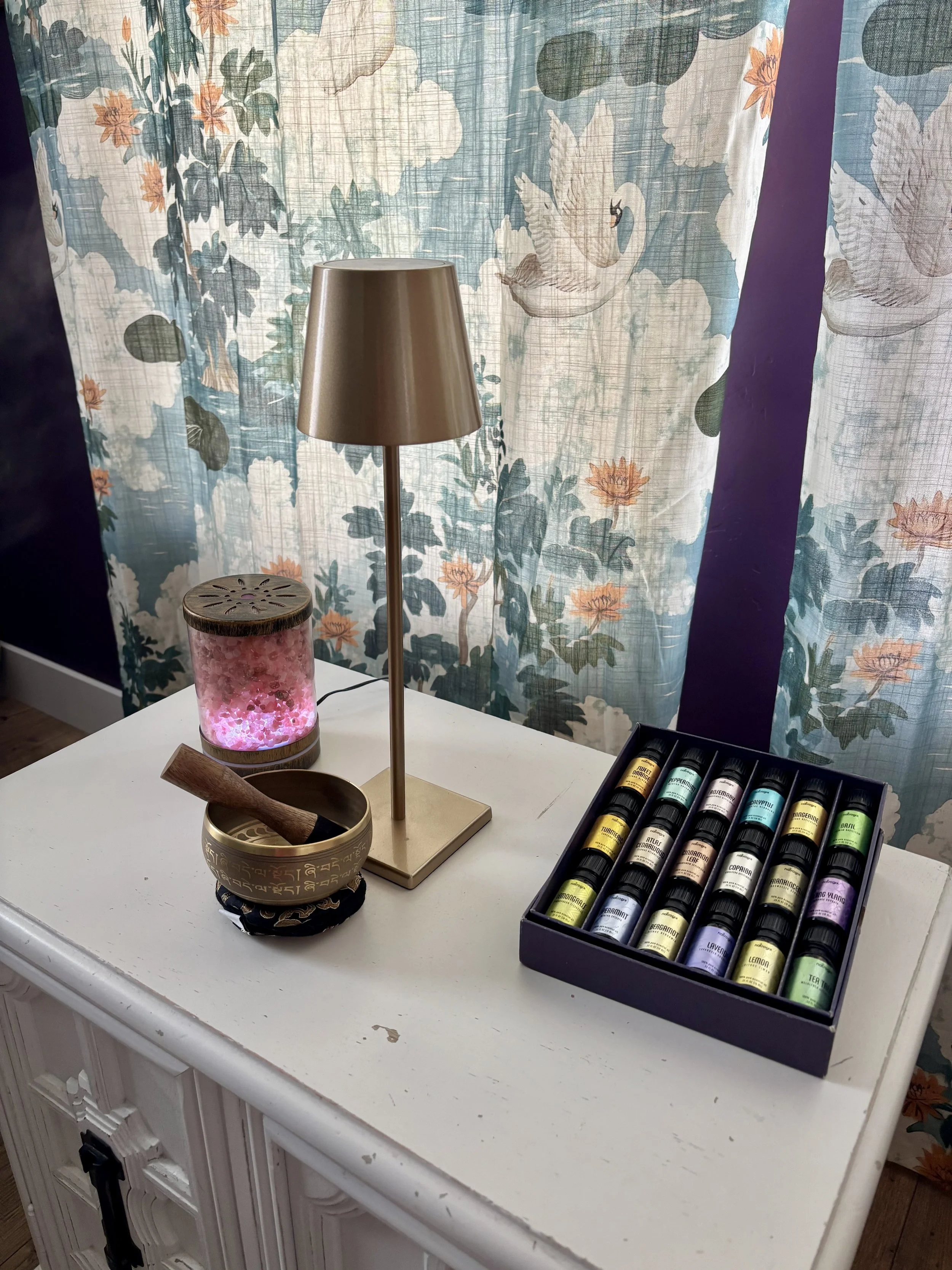 A white sideboard with a small research lamp, a box of colorful essential oils, and a mineral diffuser. Behind them are floral curtains with swan illustrations, purple accent wall, and wood flooring.