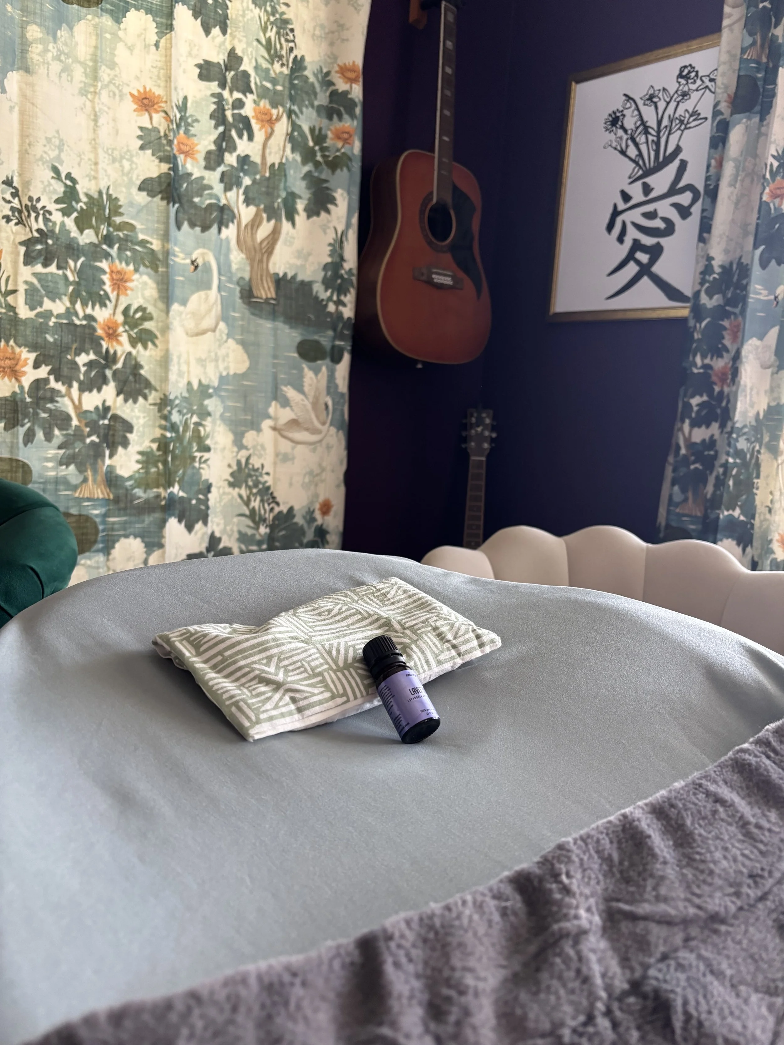 Close-up view of a bed with a beige blanket, a small towel, and a small bottle with a purple label on top. In the background, a room with a blue wall, a guitar hanging on the wall, floral curtains, and framed artwork can be seen.