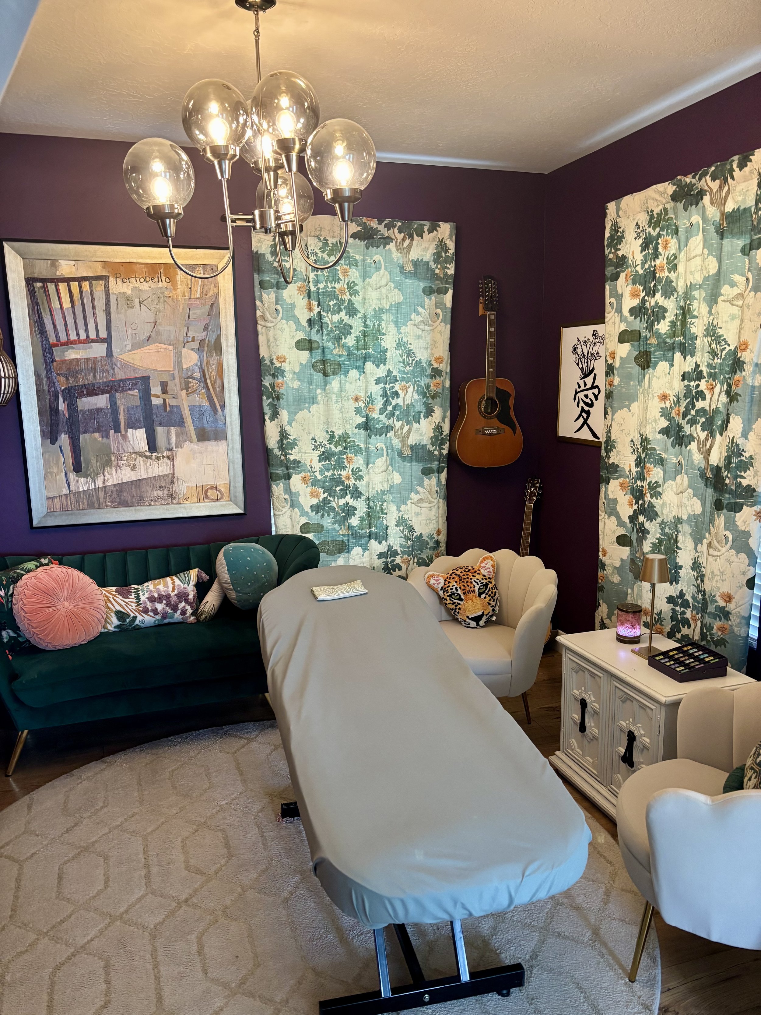 A cozy living room with a dark purple wall, floral curtains, and various decorated furniture. There is a green velvet sofa with colorful throw pillows, a white armchair with a leopard head pillow, and a round table covered with a white cloth. A modern chandelier with multiple light bulbs hangs from the ceiling, and a guitar is mounted on the wall. A painting of chairs and a framed artwork with floral text are also visible, along with a small white cabinet with a box of art supplies, a lamp, and a jewelry box.