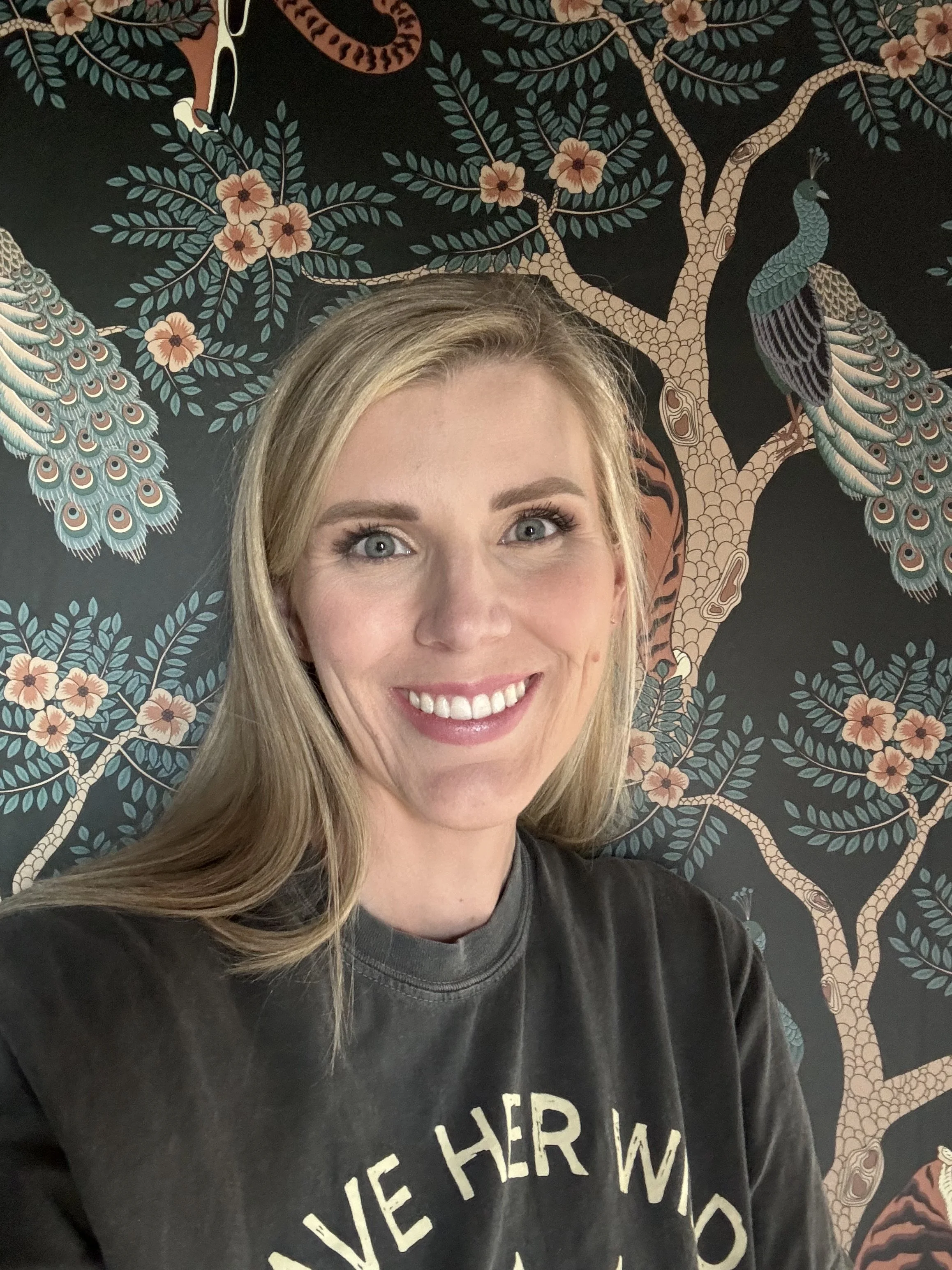 Close-up of a smiling woman with blonde hair standing in front of a decorative mural featuring a peacock perched on a tree with pink flowers against a black background.
