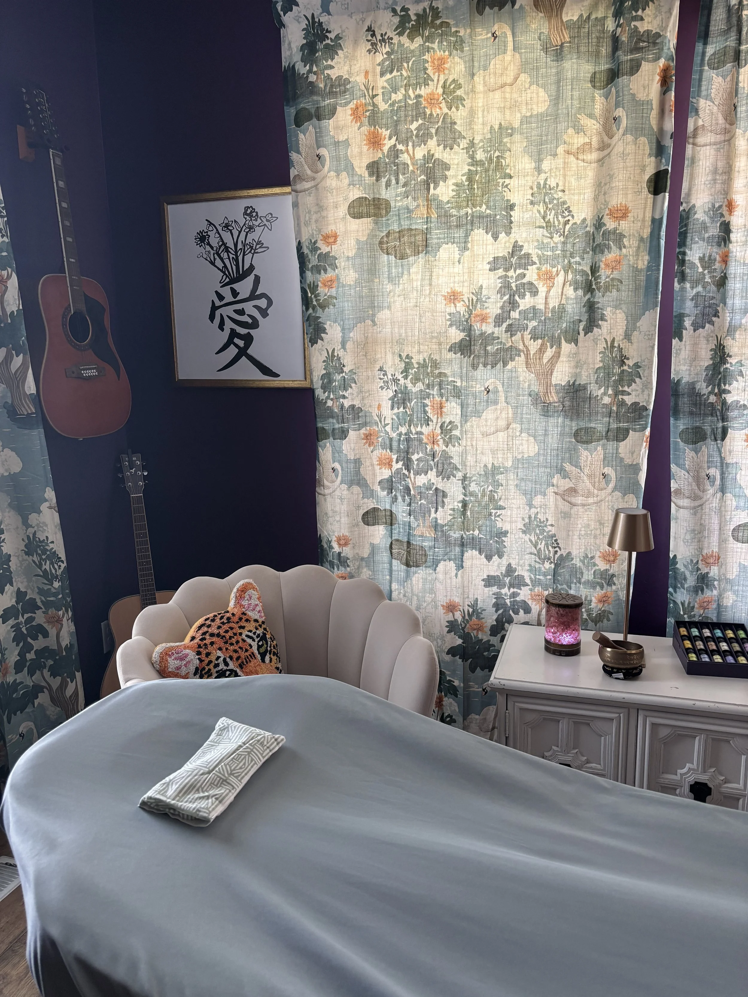 A cozy bedroom with a bed covered in a light gray bedspread, a white armchair with a cheetah-print pillow, a white side table with a pink lamp and some skincare products, and a window with floral curtains featuring swans and cherry blossoms. There are two guitars mounted on the purple wall and a framed artwork with Asian characters next to the window.