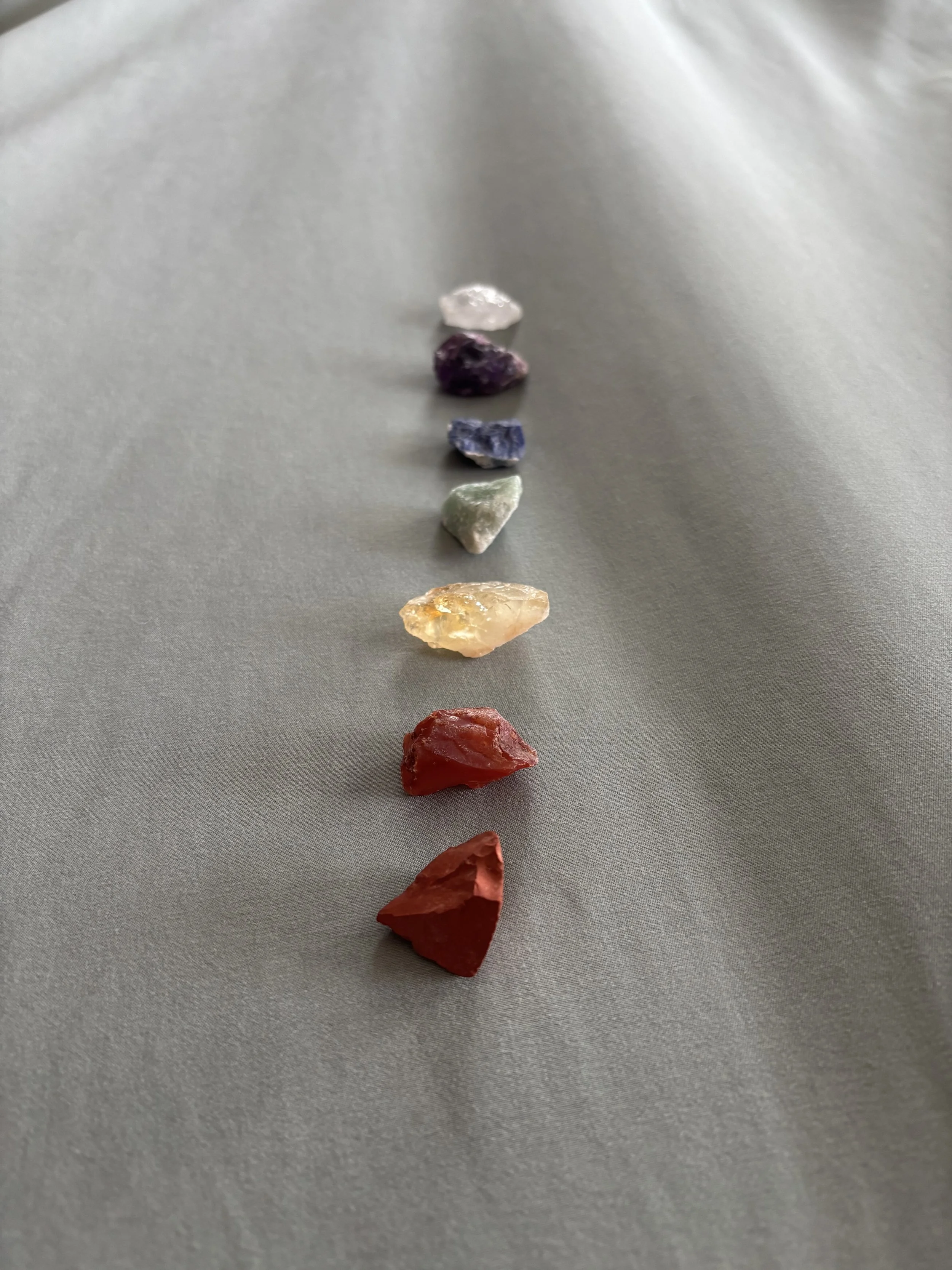 A row of seven small, raw gemstones or mineral samples in various colors, arranged in a line on a plain, light-colored surface.
