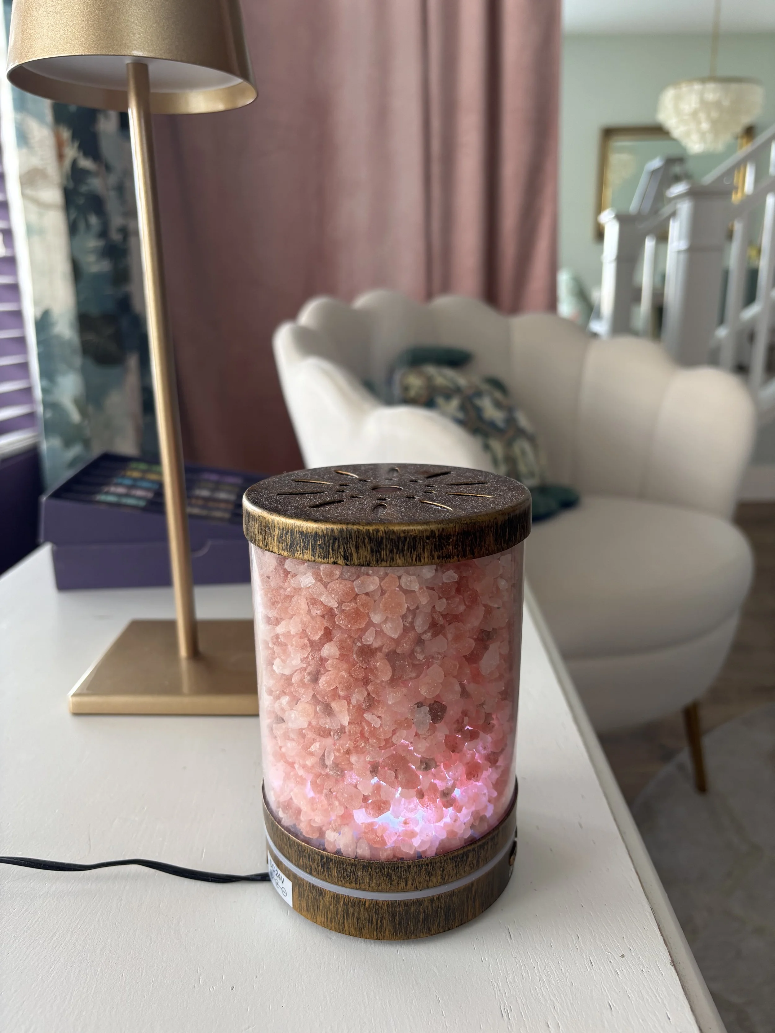A decorative lamp with a wooden base and top, and a cylindrical glass middle filled with pink Himalayan salt crystals, sitting on a white surface with a black power cord visible.