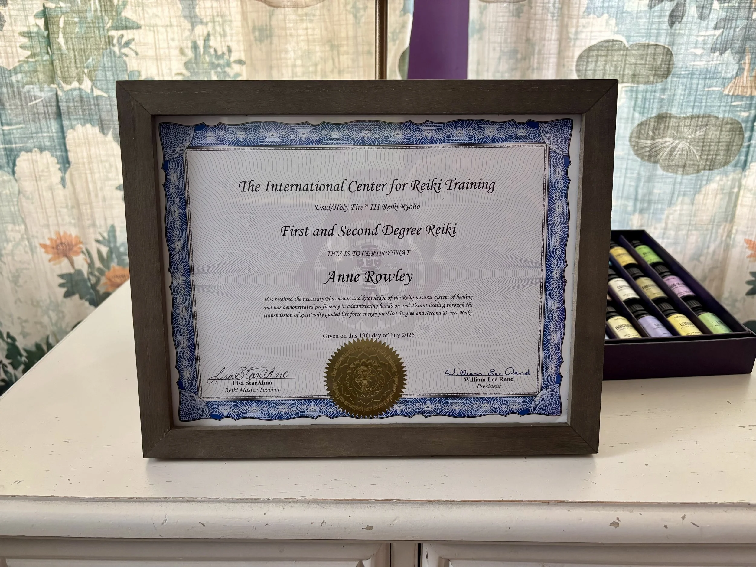 A framed certificate awarded by The International Center for Reiki Training, certifying First and Second Degree Reiki to Anne Rowley, with signatures from Lisa Star Ahna and William Lee Rand, displayed on a white surface with a background of floral and cloud-patterned curtains, and a box of essential oils nearby.