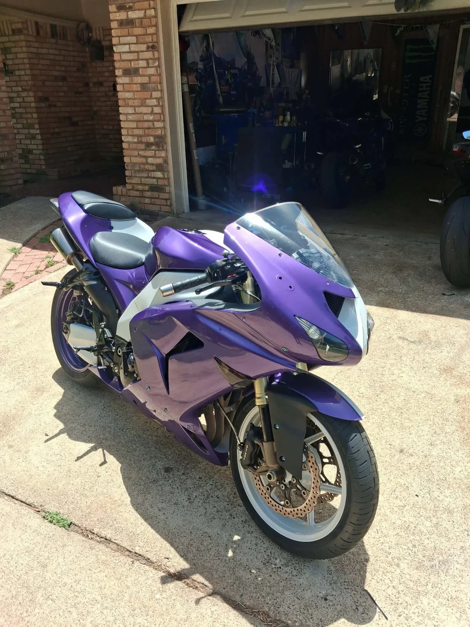 A purple sport motorcycle parked outside on a concrete driveway in front of a brick house. The garage is open and inside there are various tools and equipment, including a Yamaha banner visible on the wall.