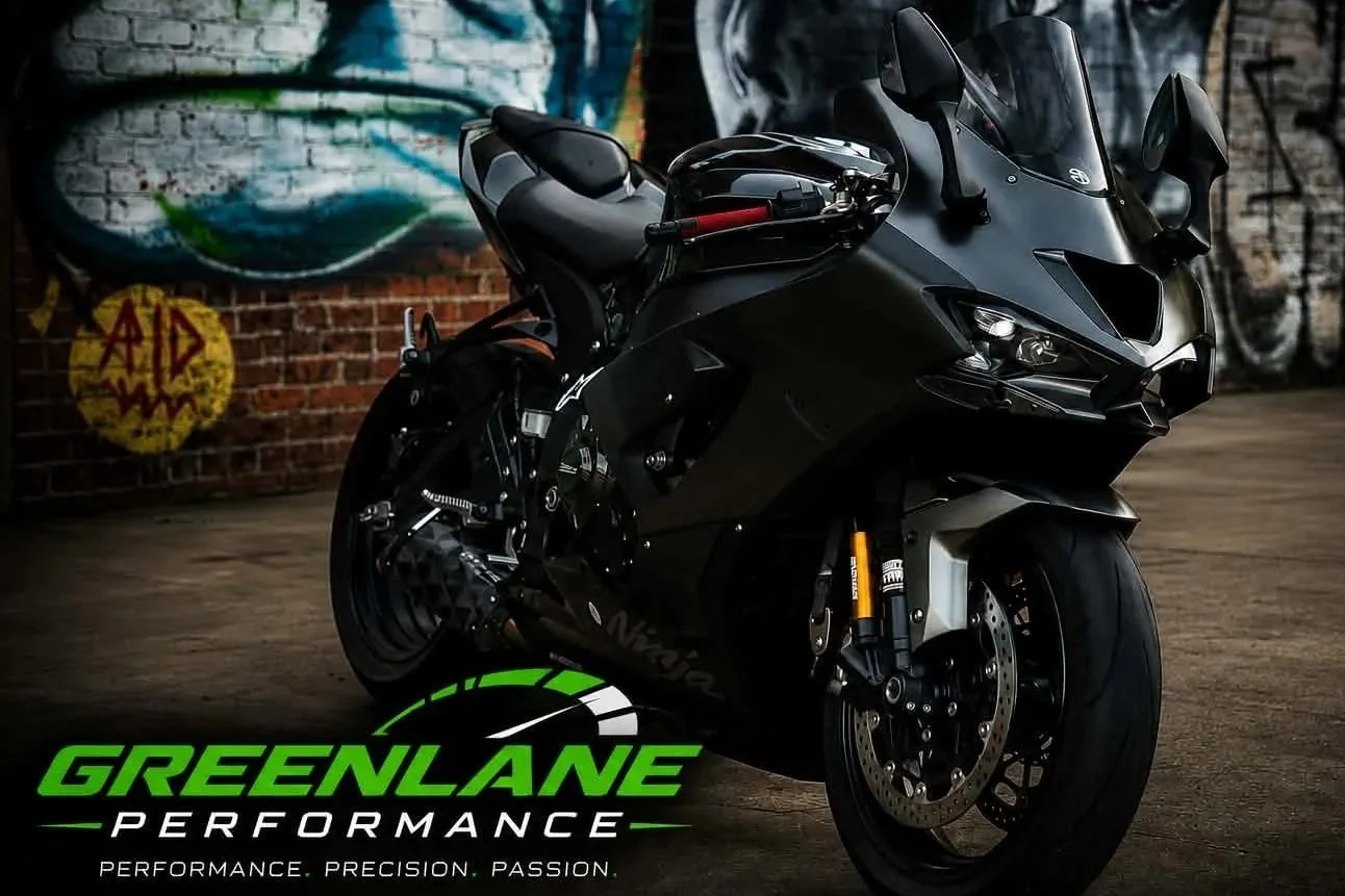 A black Kawasaki Ninja motorcycle parked indoors with a graffiti wall in the background displaying the words 'Aid' in a yellow circle, and the logo and text 'GREENLANE PERFORMANCE' along with the words 'Performance,' 'Precision,' and 'Passion' at the bottom.