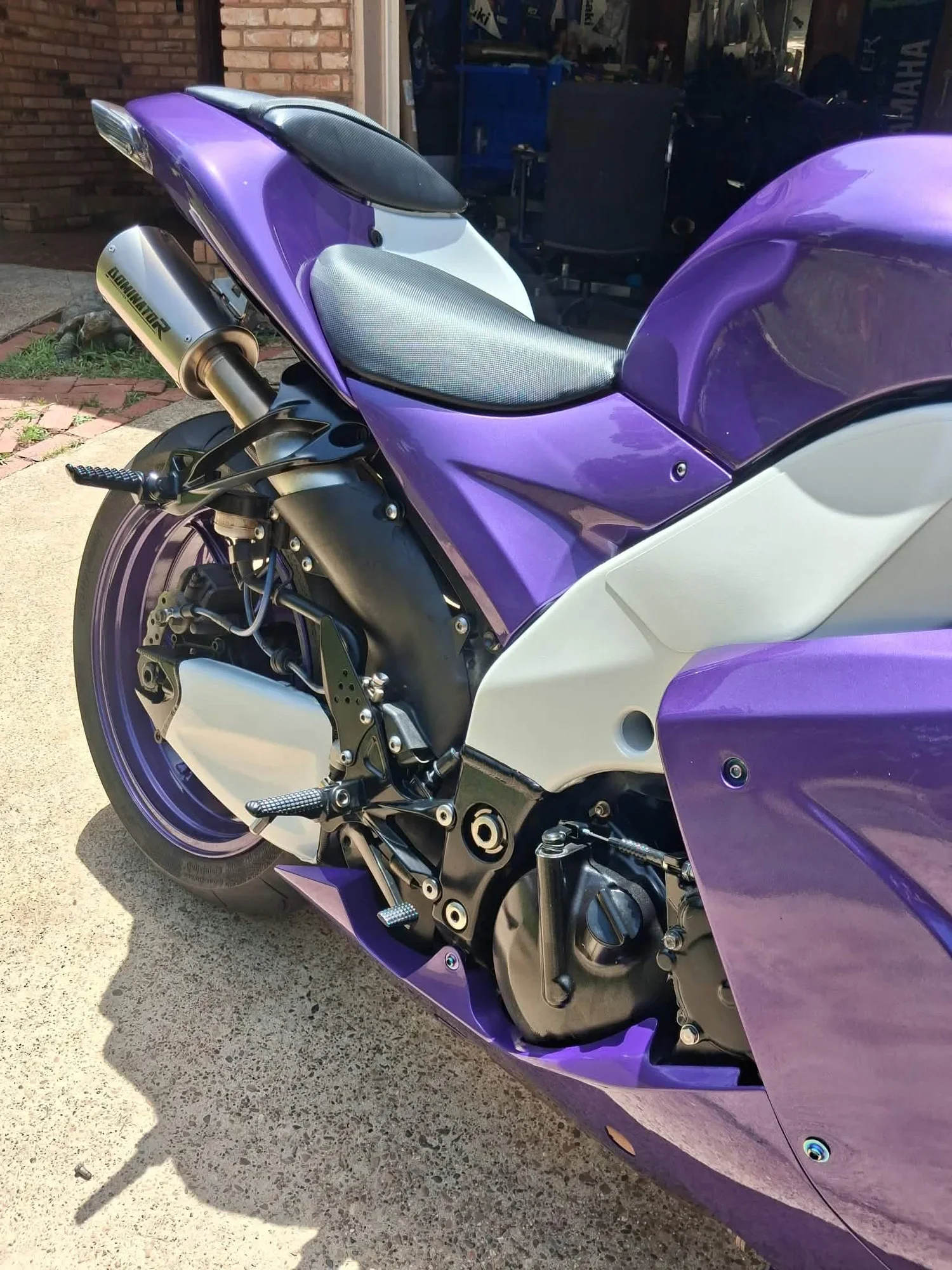 Close-up of a purple and white motorcycle parked on concrete, showing the seat, rear wheel, and exhaust system with 