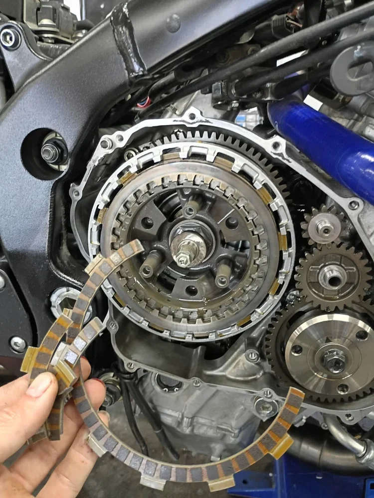 Close-up of a motorcycle engine with a mechanic holding a multi-colored clutch friction plate while working on the gears.