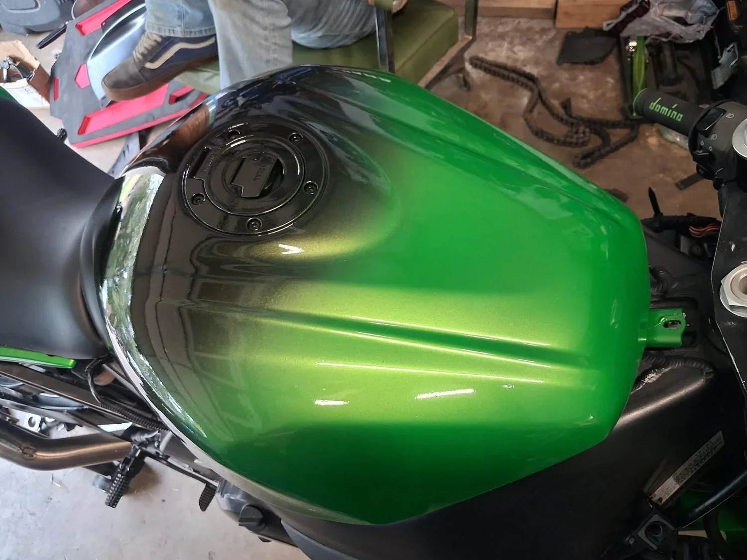 Top view of a motorcycle fuel tank painted in green with a black fuel cap and black seat, in a garage or workshop setting.