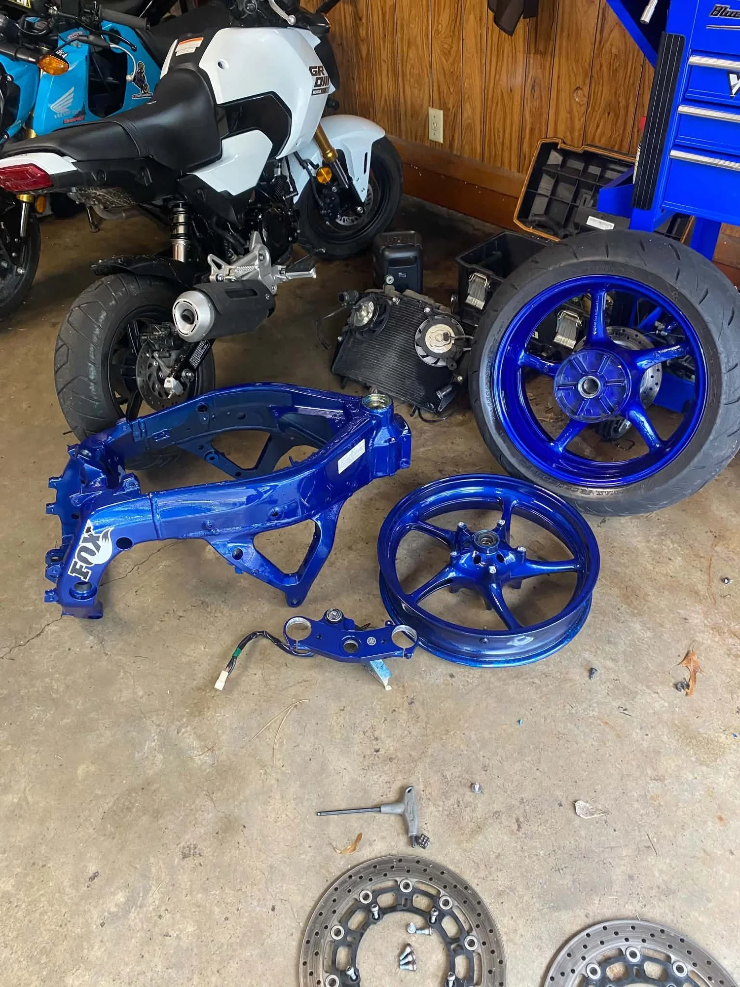 A motorcycle undergoing repairs with blue rims and various parts, including a front wheel, brake rotors, and a frame, placed on a garage floor.