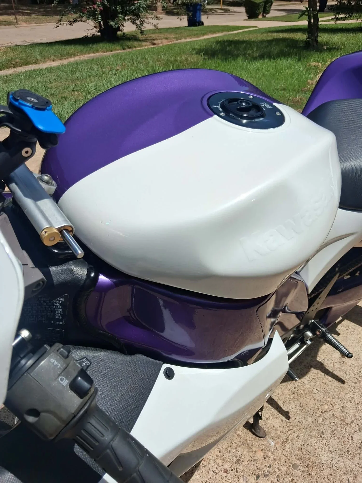 Close-up of a motorcycle's blue and white fuel tank with a black fuel cap, part of the seat, handlebars, and side stand visible, parked outdoors on a sidewalk with grass and trees in the background.