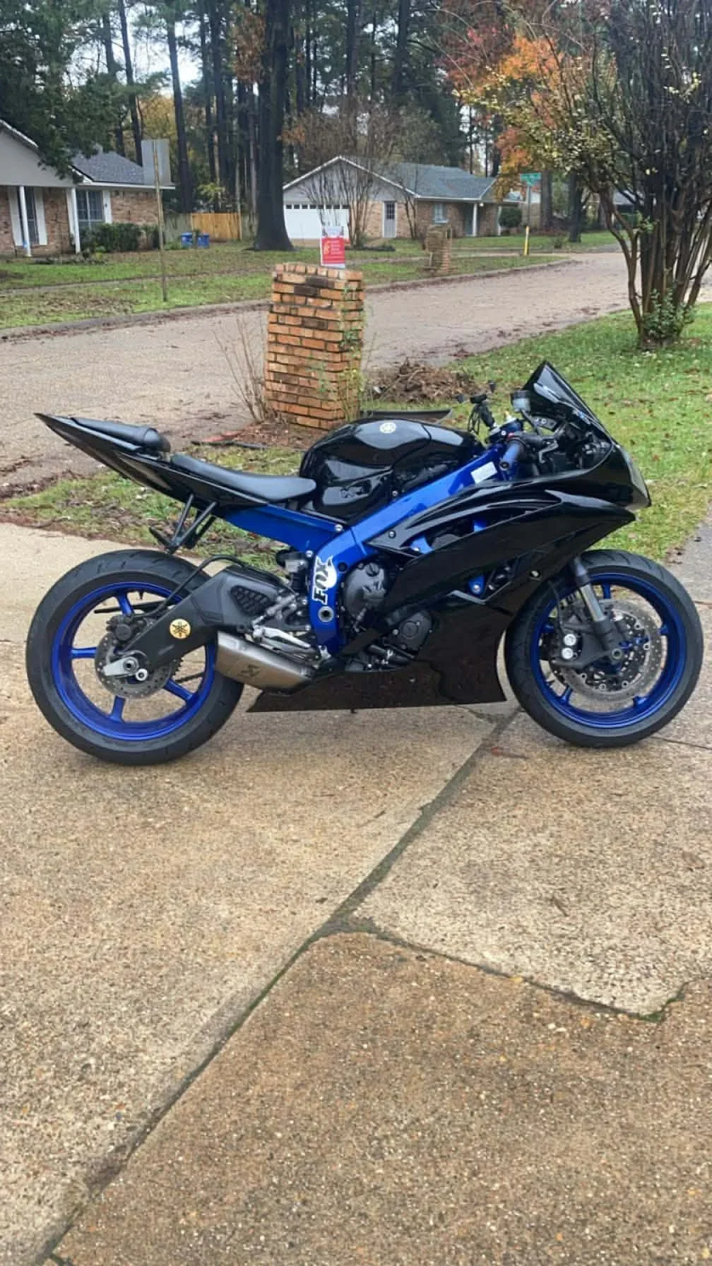 A black and blue Yamaha motorcycle parked on a concrete driveway in front of a residential neighborhood with trees, houses, and a brick mailbox.