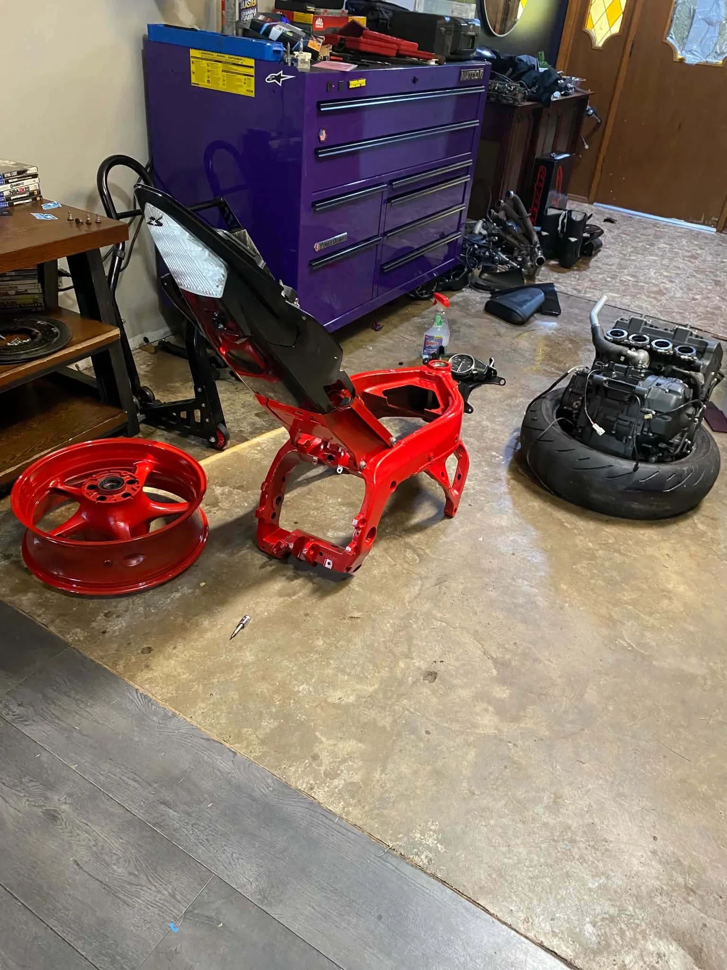 A garage workspace with a large purple toolbox, disassembled motorcycle parts including red wheels, engine, and frame, a spray bottle, and a wooden workbench with DVDs and small items.