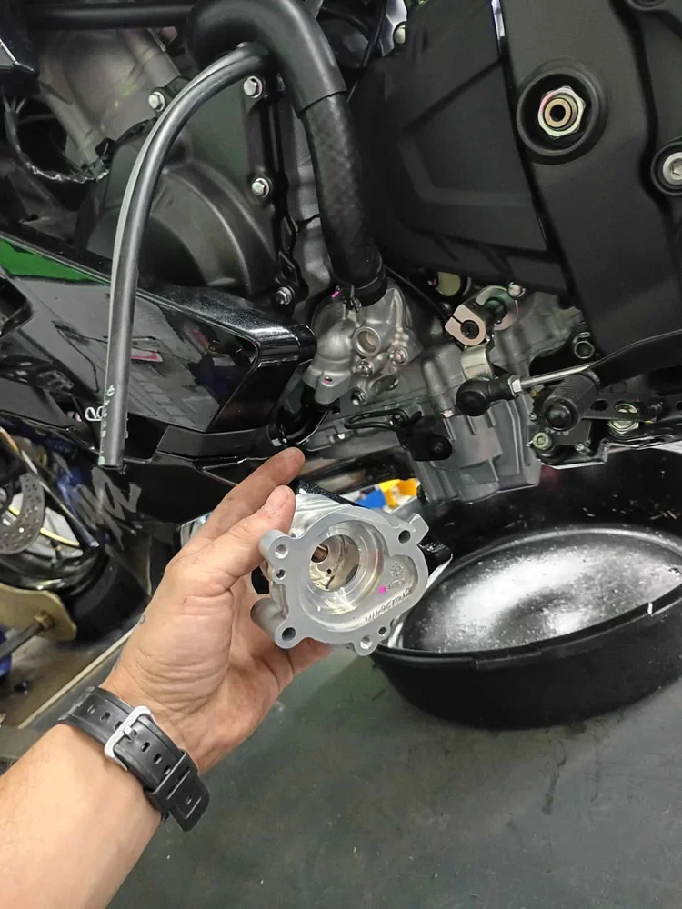 Hand holding a metal motorcycle part near the engine, with a black bowl on the ground nearby.