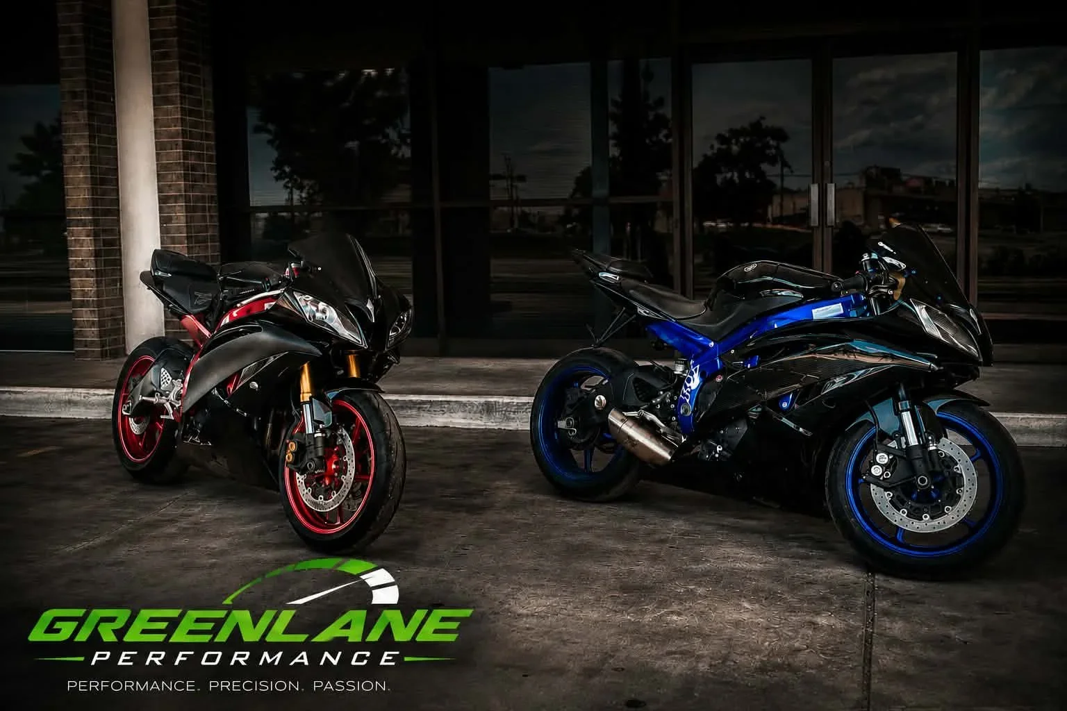 Two sport motorcycles parked outside a building at night, one black and red, the other black and blue, with Green Lane Performance logo and slogan at the bottom.