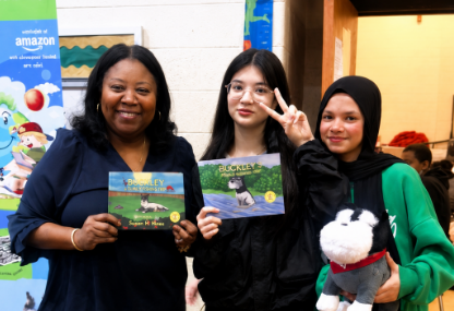 Three women standing indoors, holding children's books and a stuffed animal, smiling for the photo. The woman on the left holds a book, the woman in the middle makes a peace sign, and the woman on the right holds a stuffed animal. A poster and some background people are visible.