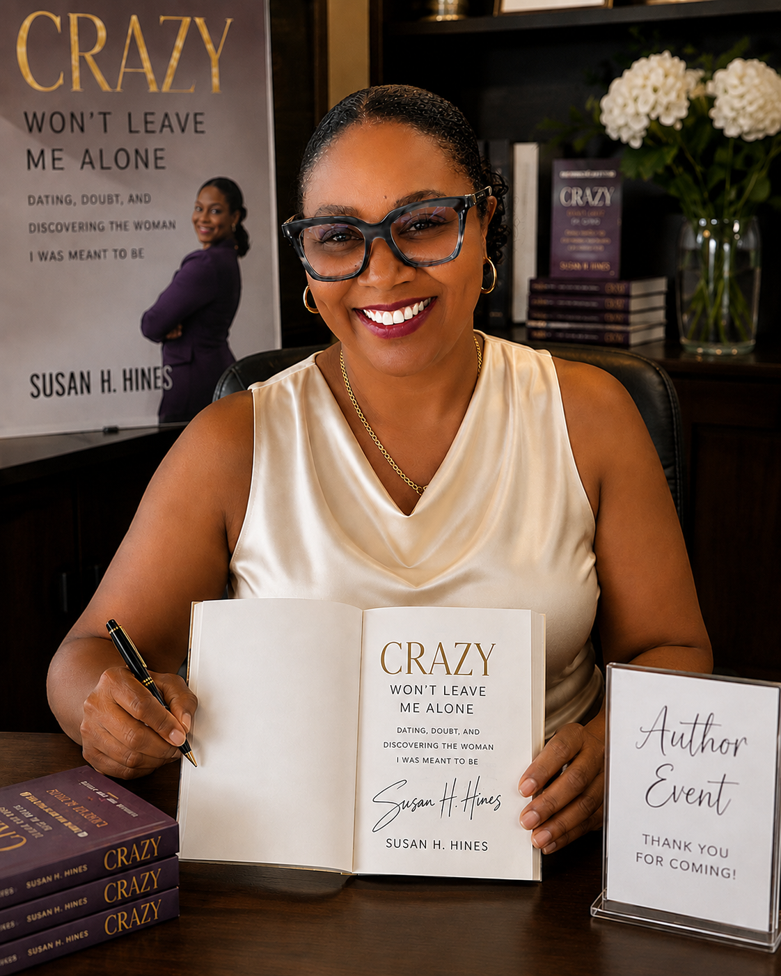 A woman at a book signing event holding a copy of her book titled 'Crazy Won't Leave Me Alone' by Susan H. Hines. She is smiling, wearing glasses, a cream-colored satin top, gold jewelry, and sitting at a table with copies of her book and a sign thanking attendees.