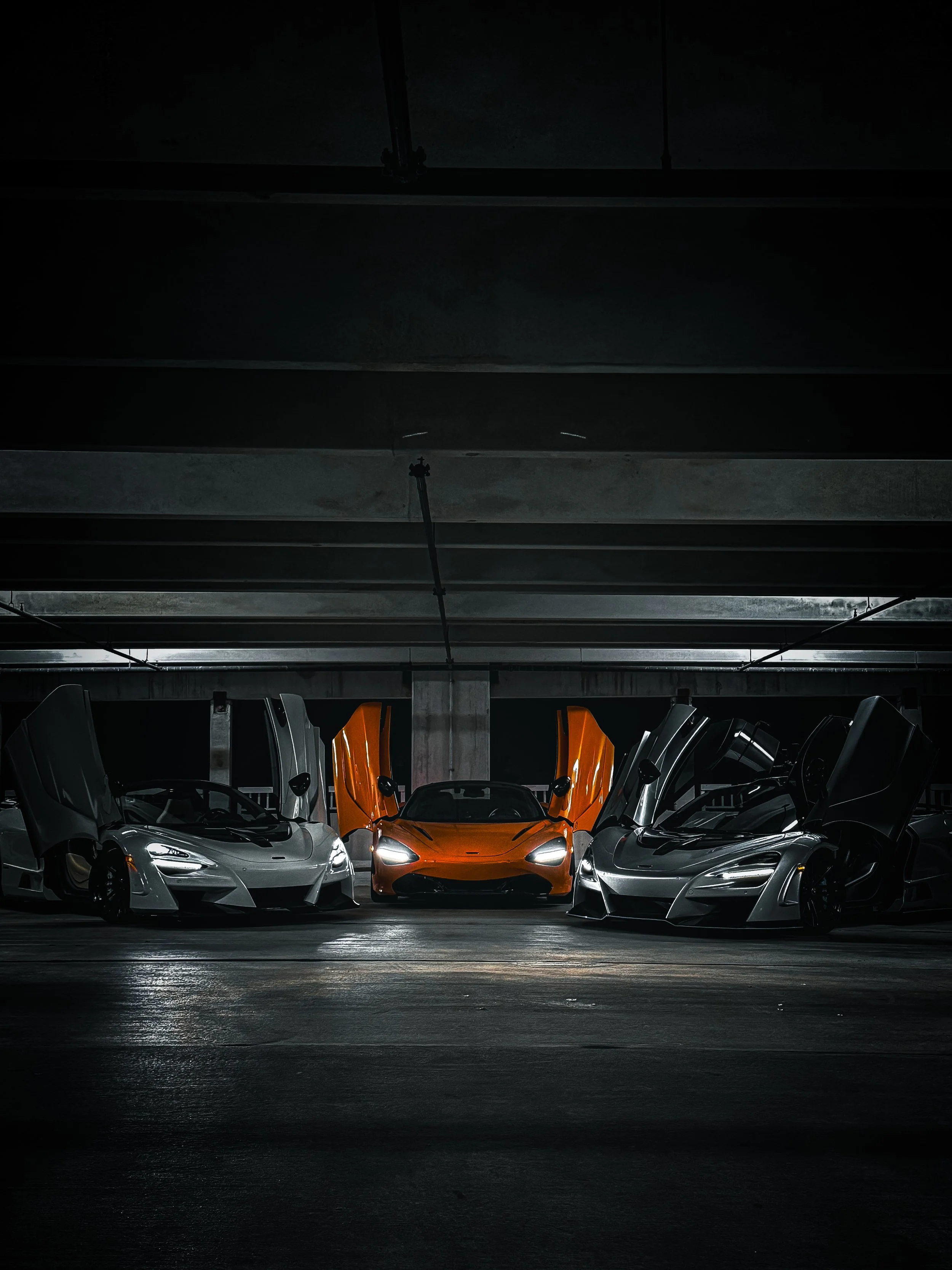 A bright orange sports car with its doors open, flanked by silver sports cars with closed doors, in a dark parking garage.