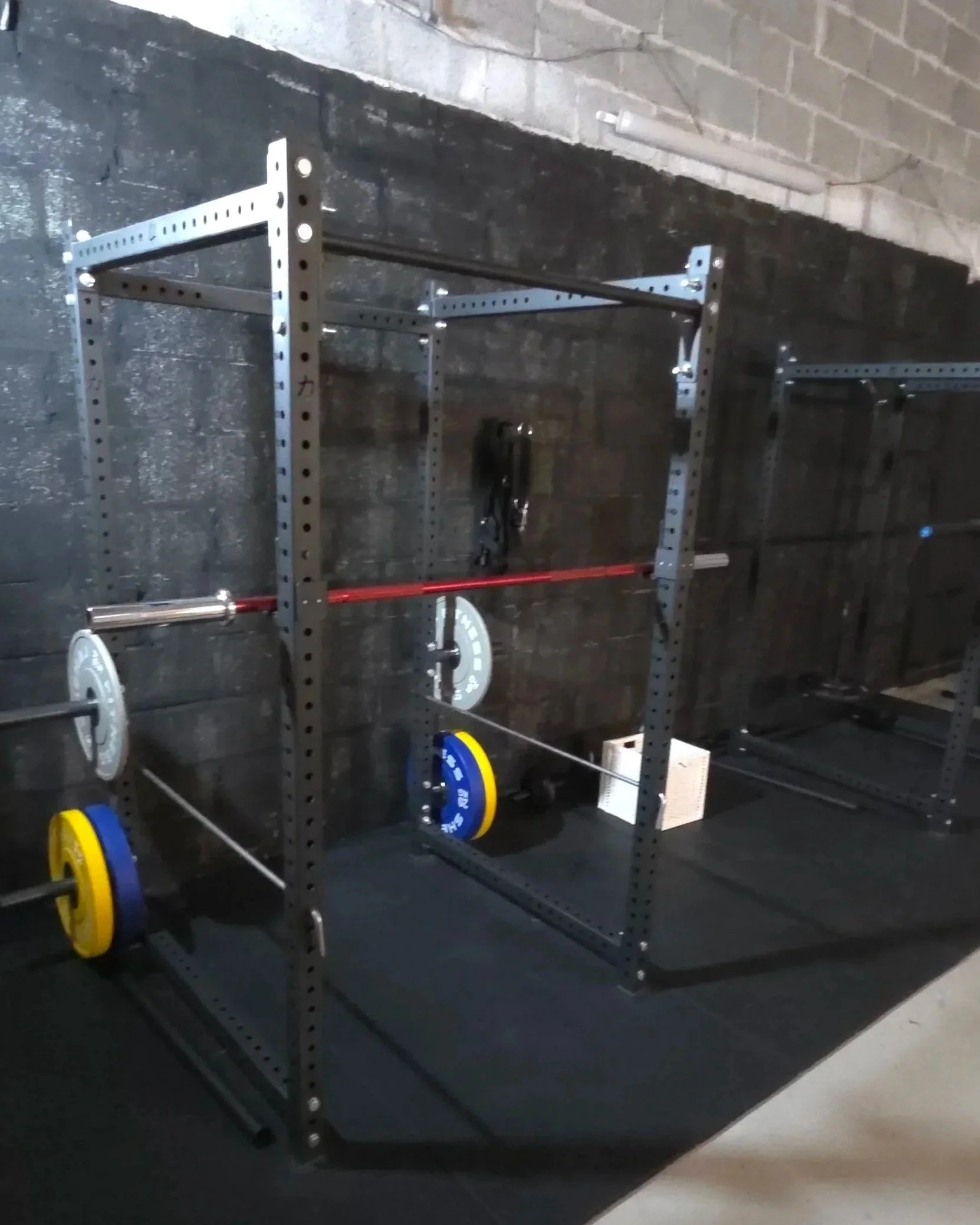 A metal fitness rack with weight plates and a barbell in a gym or workout space with a brick wall background.
