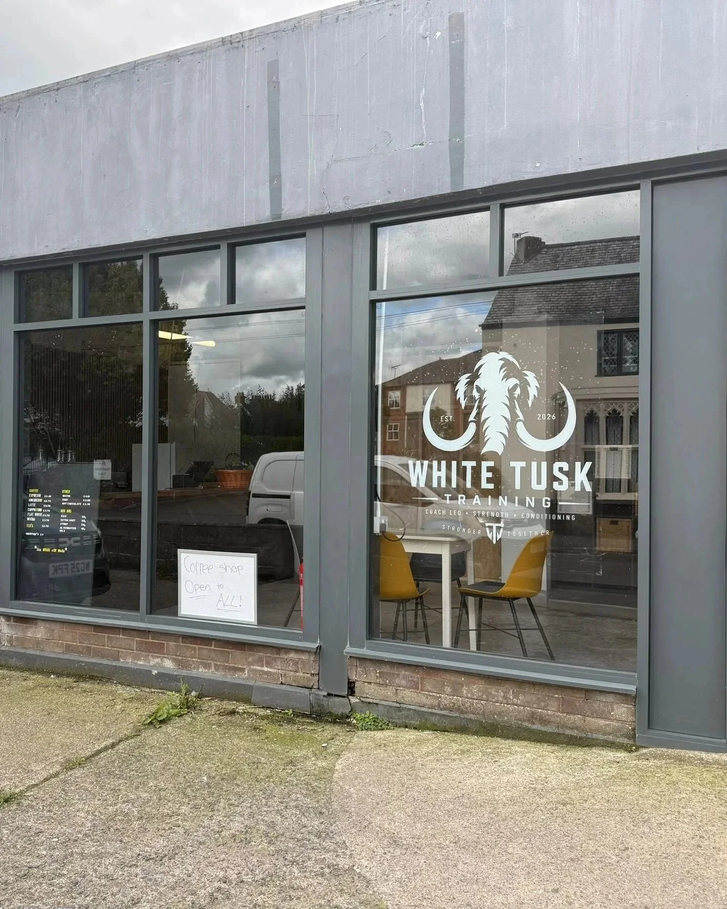 The storefront of White Tusk Training, a fitness or training center, with large glass windows showing an interior with yellow chairs. The window has white branding with a Tusks logo and text that says 'EST. 2026 White Tusk Training Coach Leo - Strength - Conditioning.' A handwritten sign on the window states 'Coffee shop open to all!'