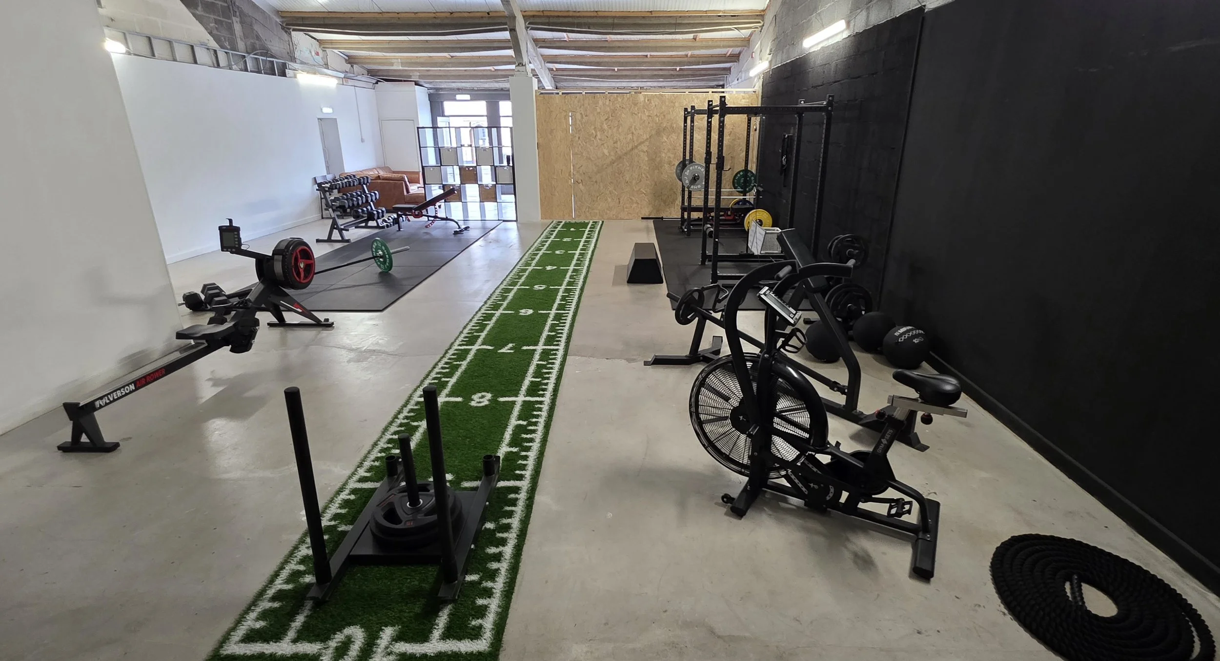 Fitness gym with workout equipment including a rowing machine, weightlifting barbell with weights, a punching bag, and a running bike. There is a green artificial turf strip with a football field design on the floor, and a seating area with chairs and a couch near a window and a decorative room divider.