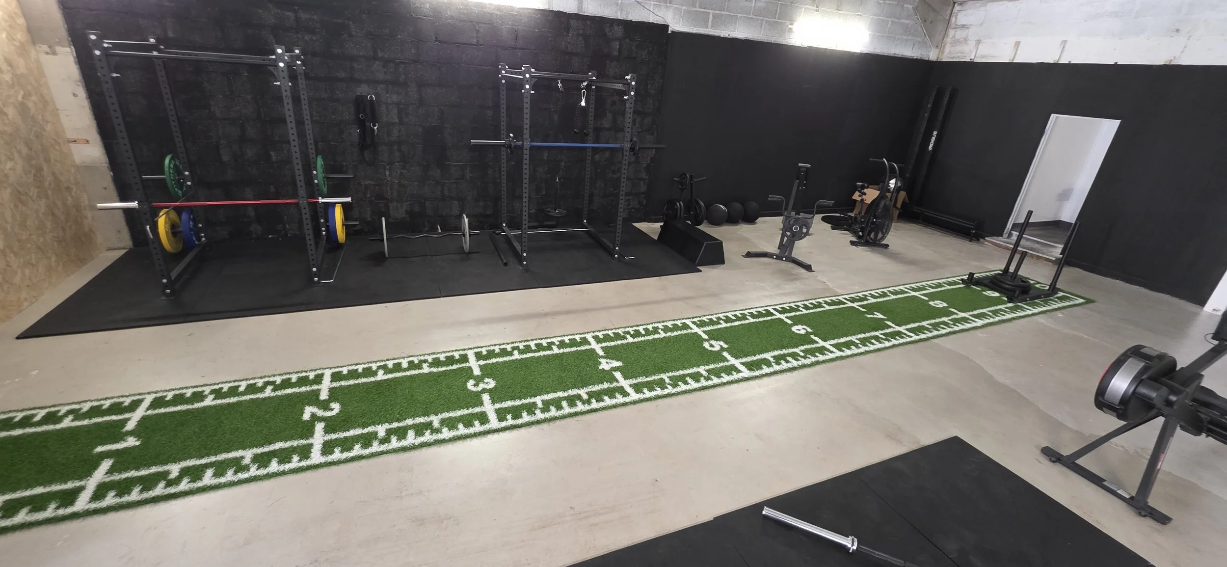 Home gym setup with weightlifting equipment, a rowing machine, exercise bike, and footprinted green turf for sprints or agility drills, inside a room with black and white walls.