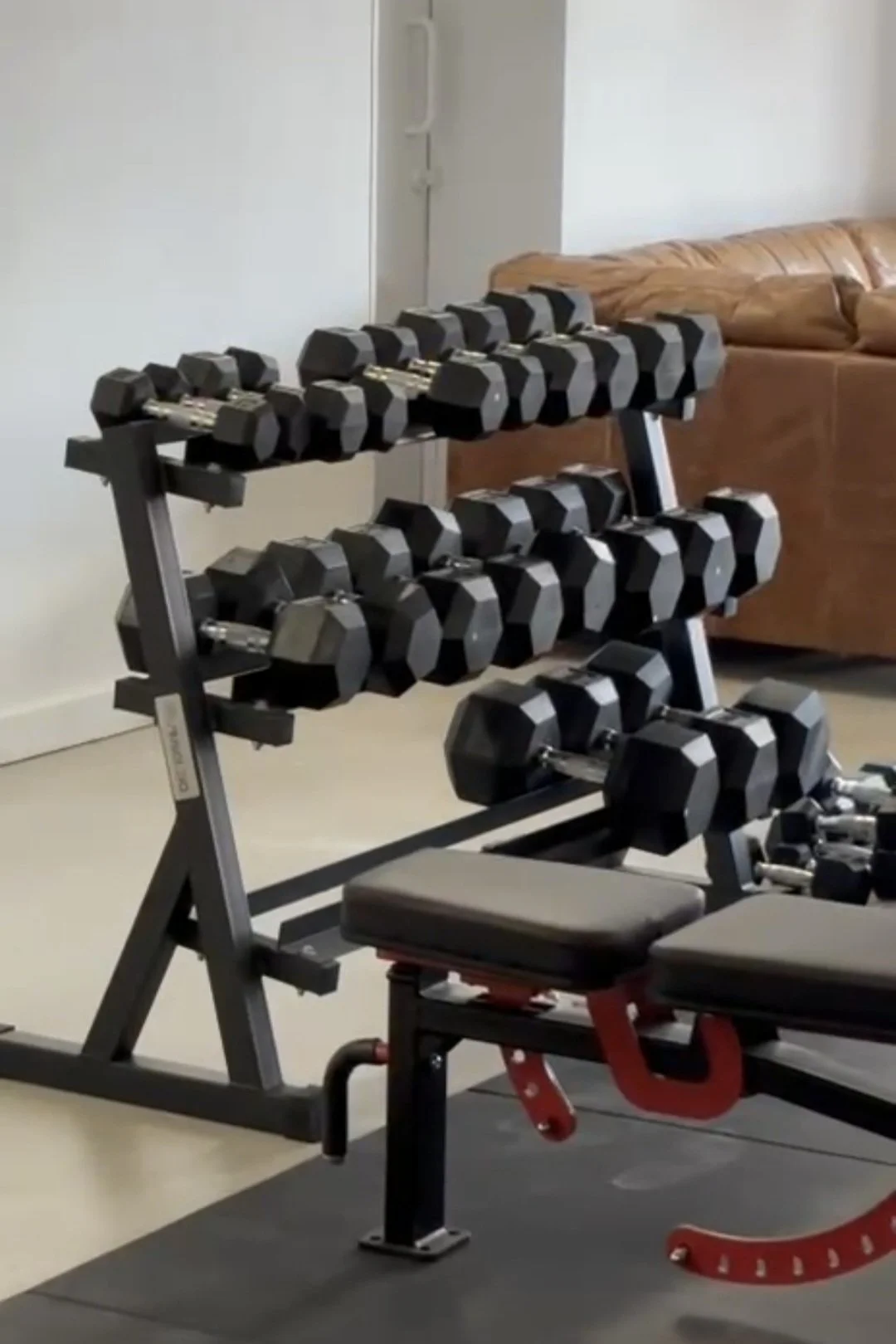 Dumbbell rack with multiple black dumbbells in a home gym with a brown couch in the background.
