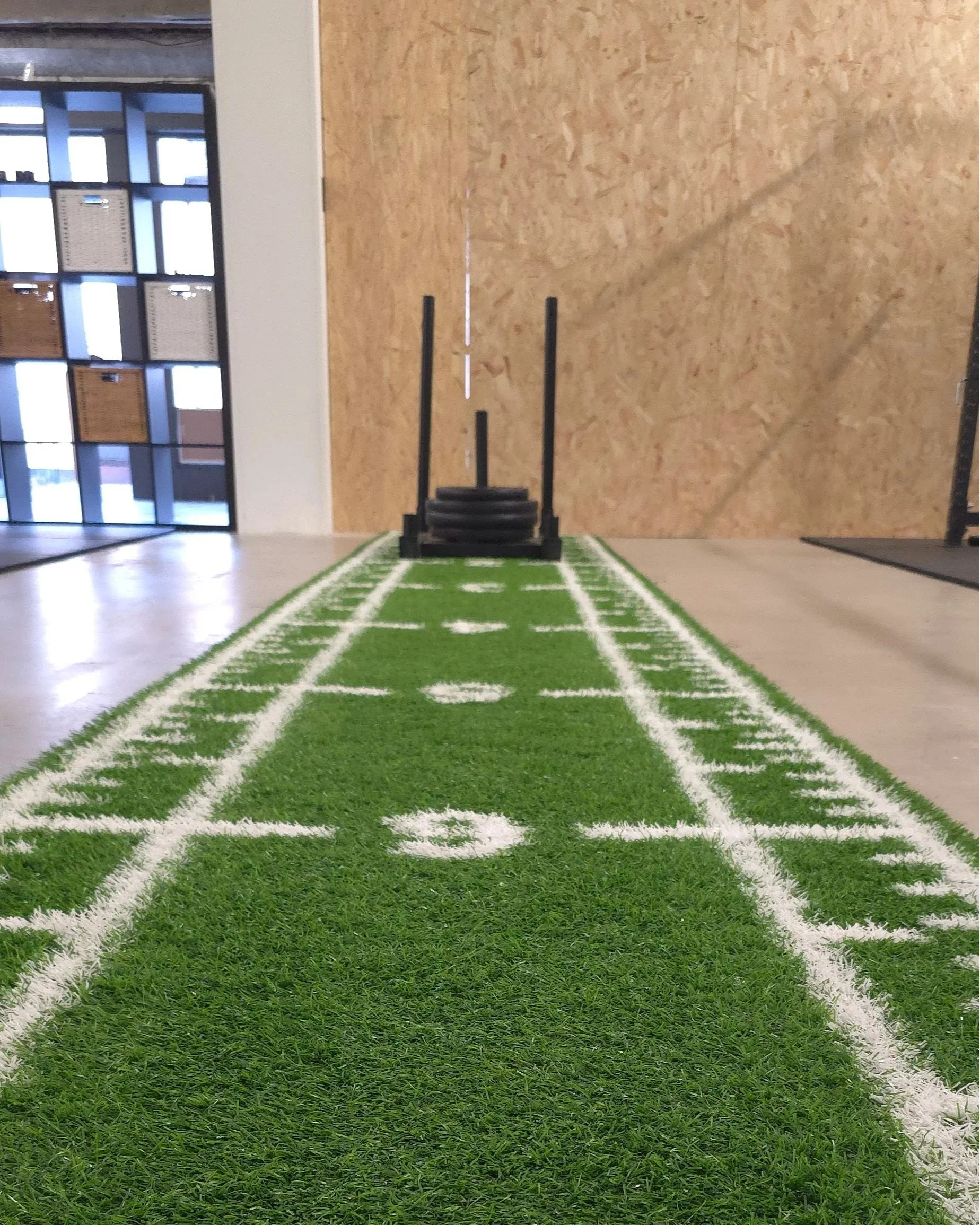 Interior of a gym with a green turf floor and a black sled with weights on it, against a wall with a plywood panel, and lockers on the left side.