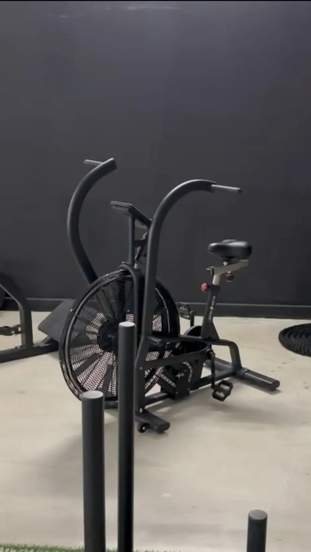 Indoor gym with a black exercise bike positioned against a dark wall, surrounded by black posts and a floor with a concrete look.