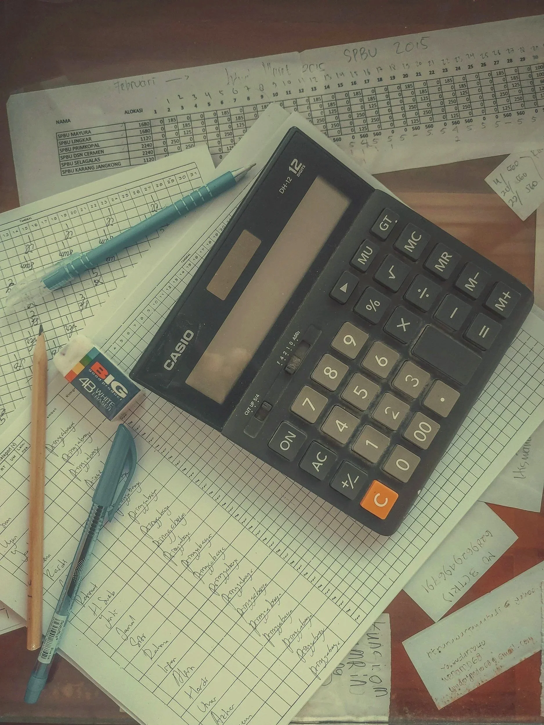 Casio desktop calculator, blue pens, pencil, and handwritten accounting worksheets with tabulated figures on a desk